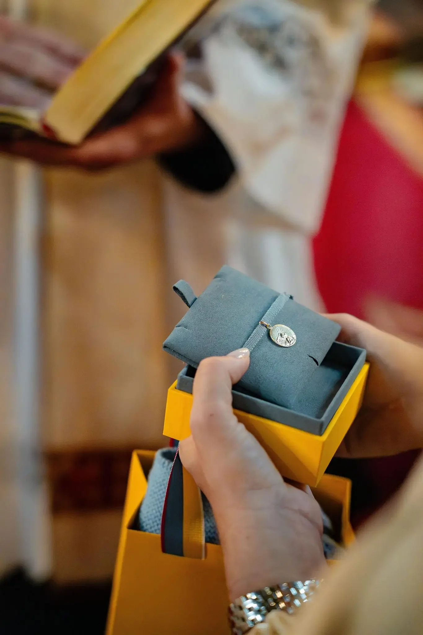Une personne tenant une boîte à bijoux avec un pendentif en or en forme de pièce ou de médaille. La personne porte une montre en métal et une robe de couleur claire. La boîte est jaune avec un intérieur gris.
