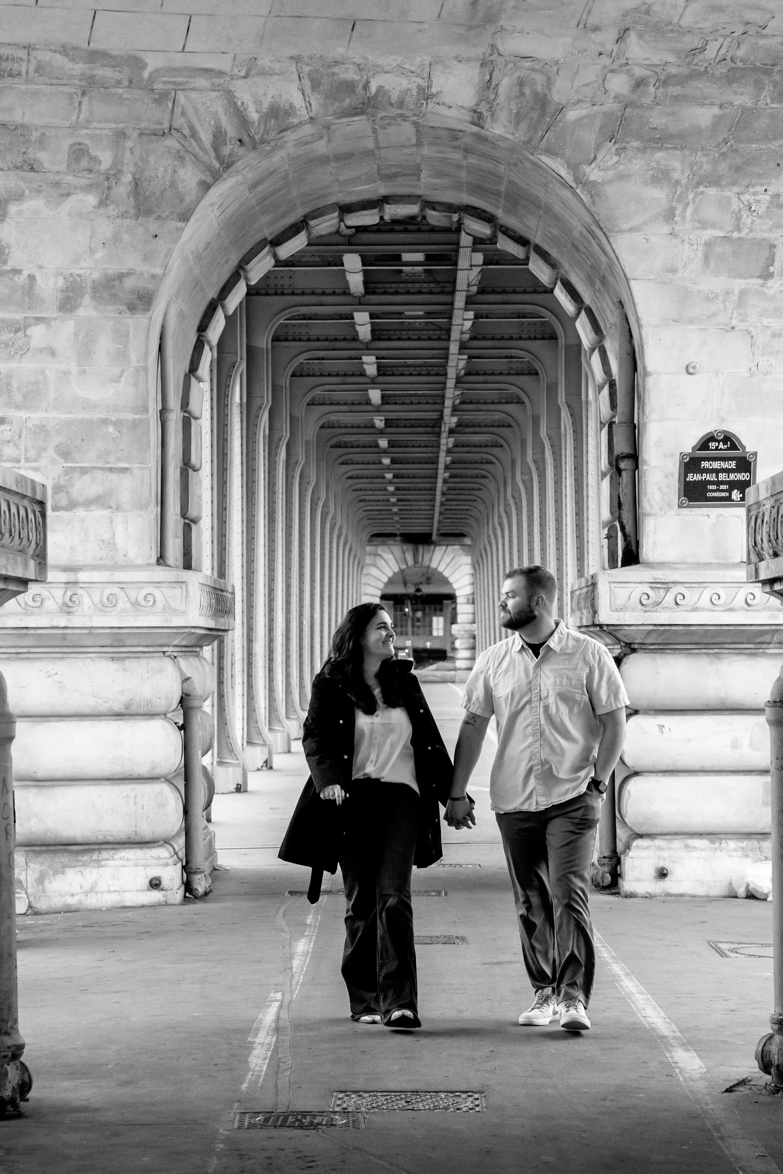Un couple marche main dans la main dans une arche en pierre sous un passage couvert, discutant et souriant.