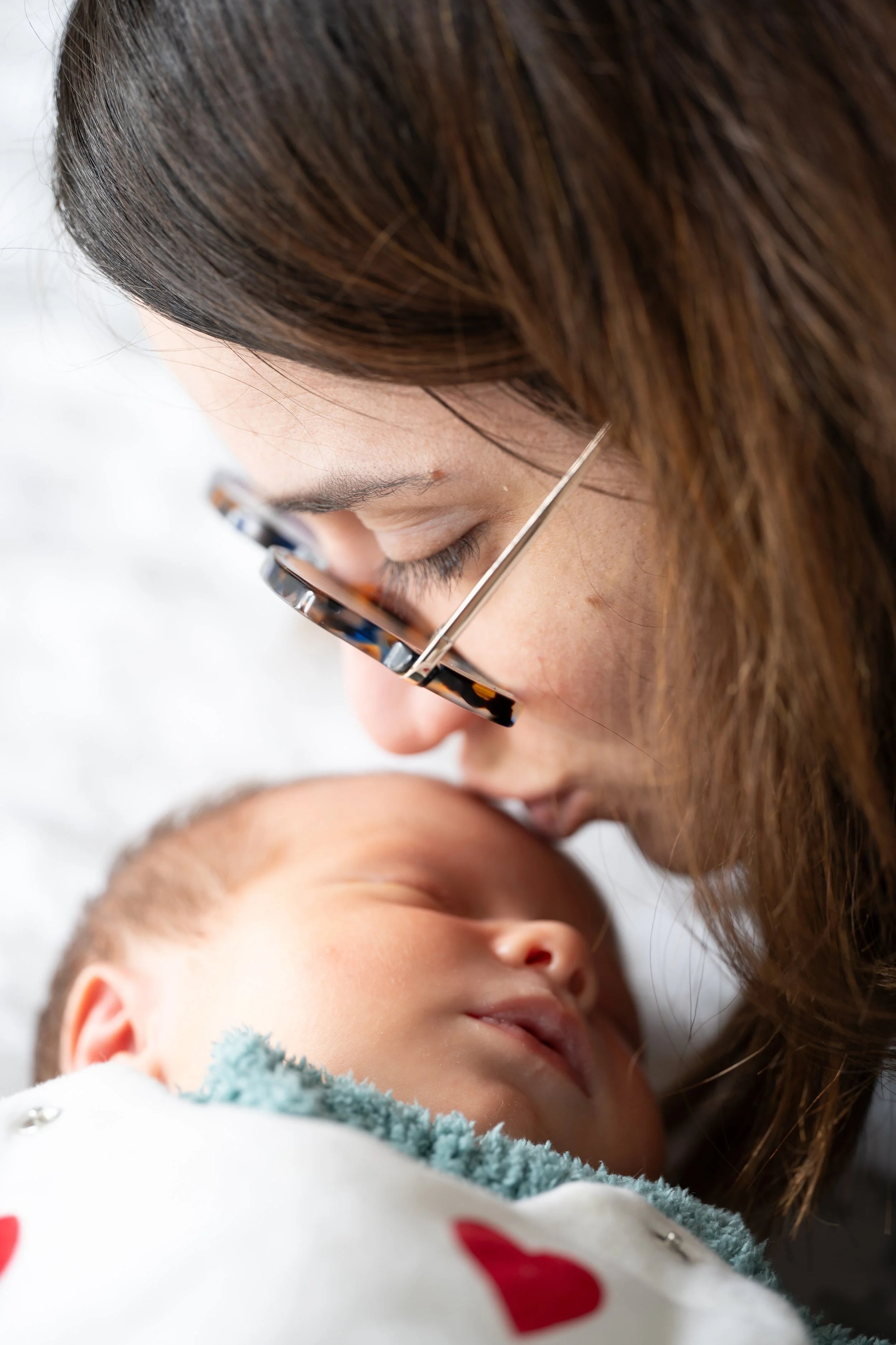 Une femme porte des lunettes de vue et regarde un bébé endormi. La femme a des cheveux bruns et longs.