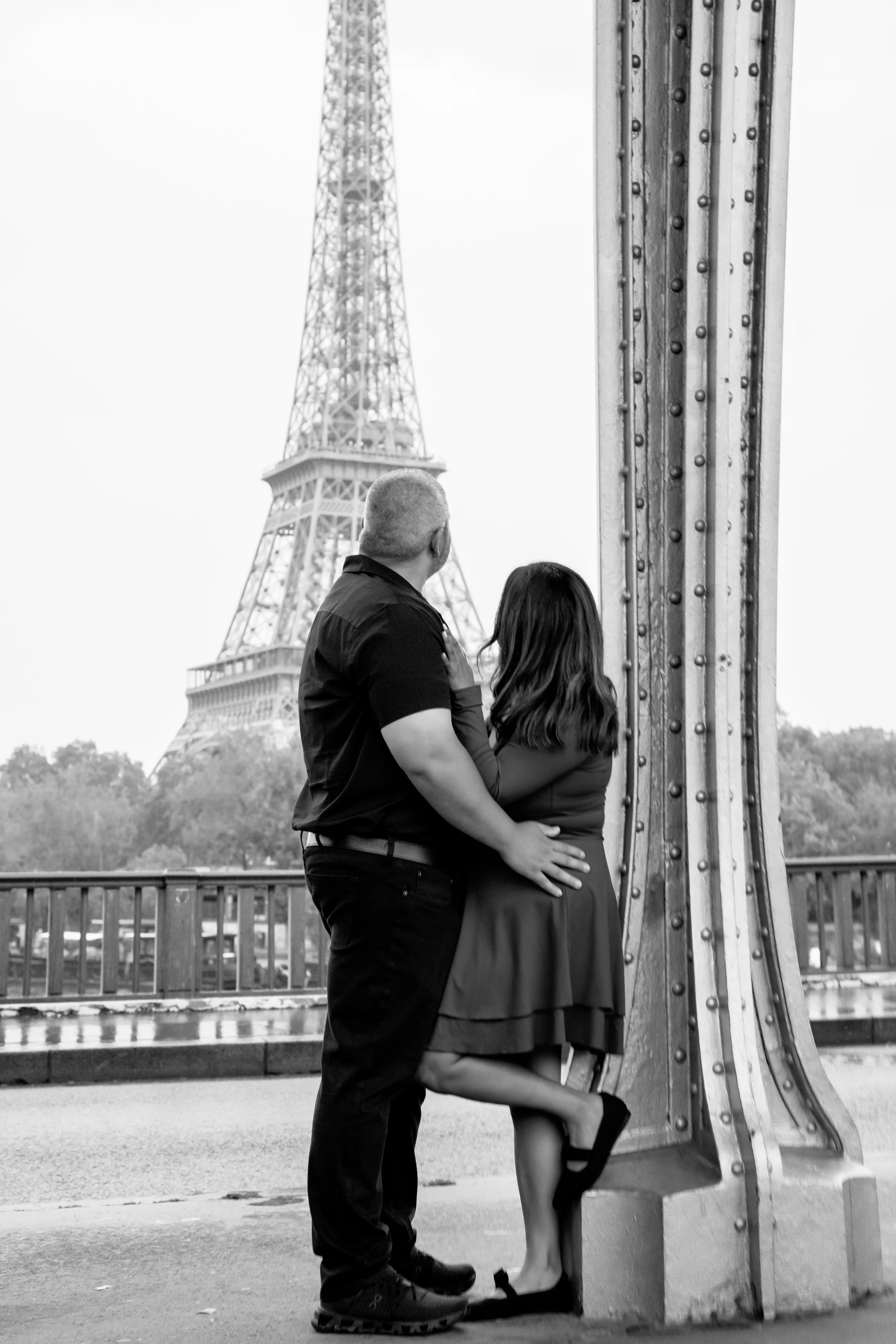 Un couple devant la Tour Eiffel, posé près d'une structure métallique. La femme est assise sur une plateforme, le homme se tient debout face à elle. La scène est en noir et blanc.