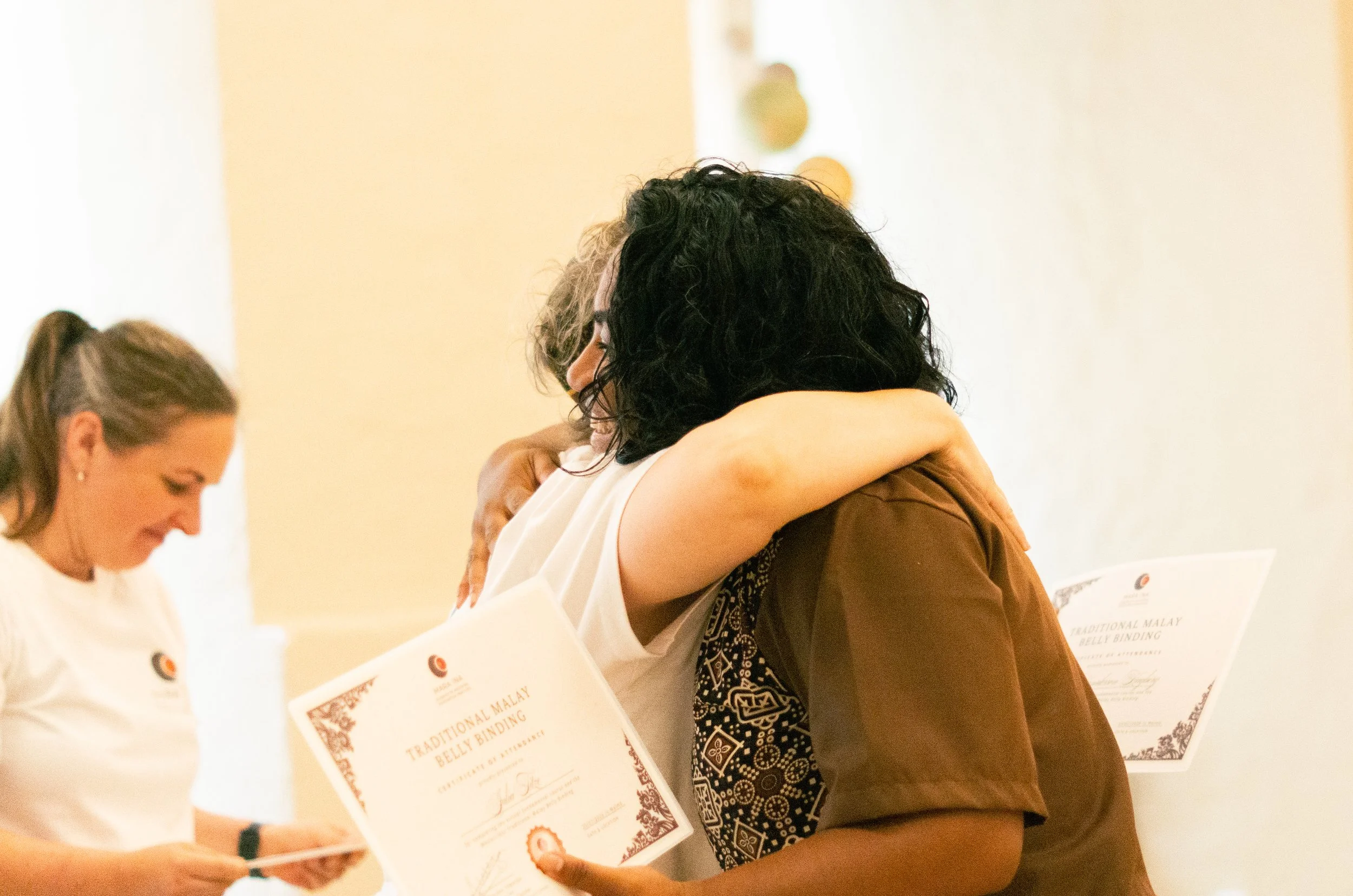 Two women hugging each other at a ceremony, holding certificates titled 'Traditional Malay Belly Binding' in a bright room.