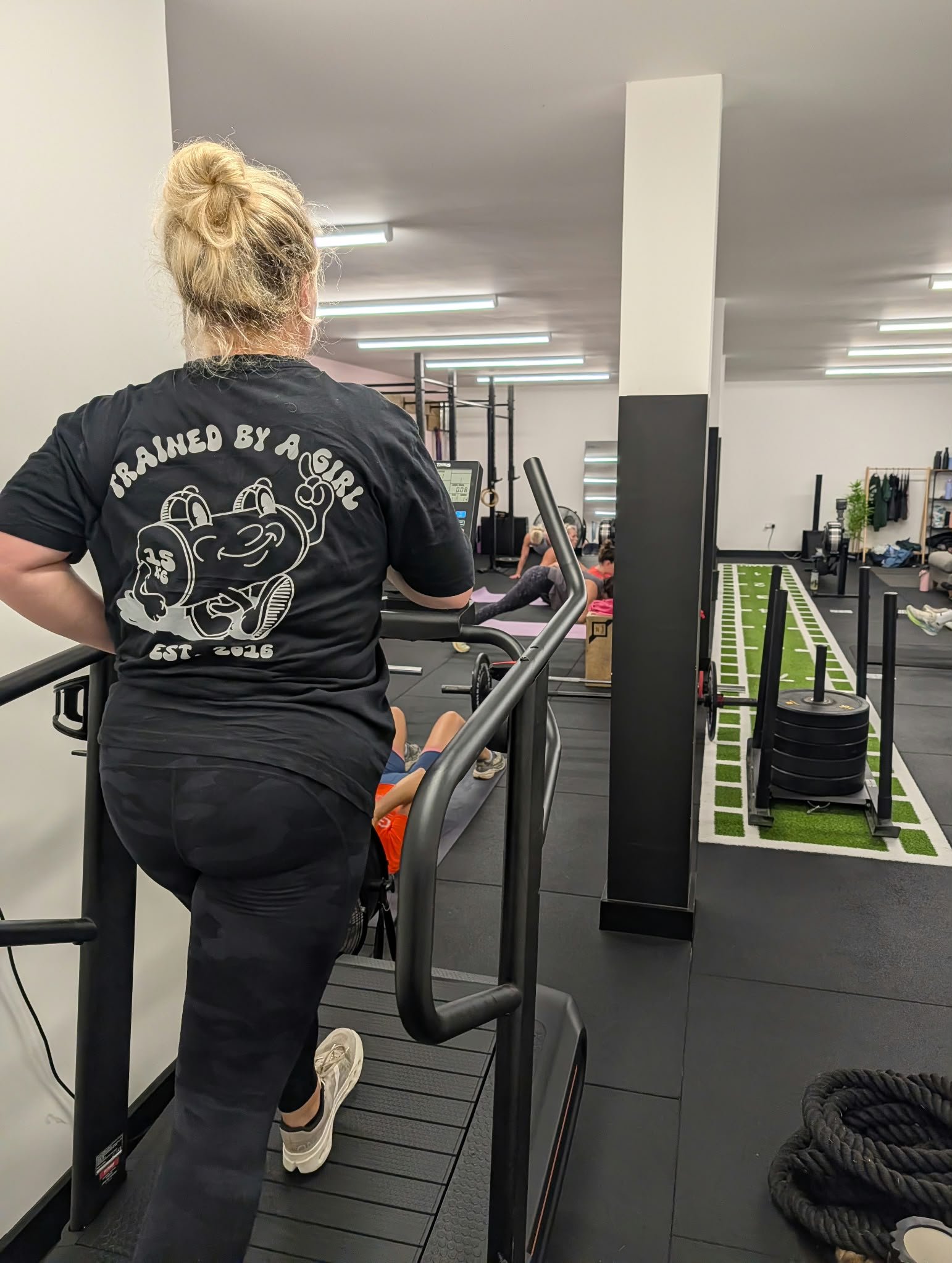 A person with blonde hair in a bun, wearing a black T-shirt that says "Trained by a Girl," is on a treadmill in a gym. The background shows people exercising, gym equipment, and a green artificial turf area.