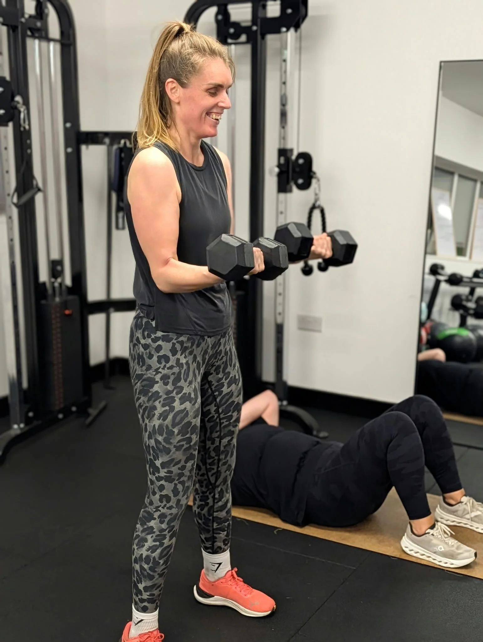 A woman with blonde hair tied in a ponytail, smiling as she lifts black dumbbells in a gym, wearing a black sleeveless top, leopard print leggings, white socks, and coral sneakers. Behind her, a person in black workout clothes is lying on a mat, and 