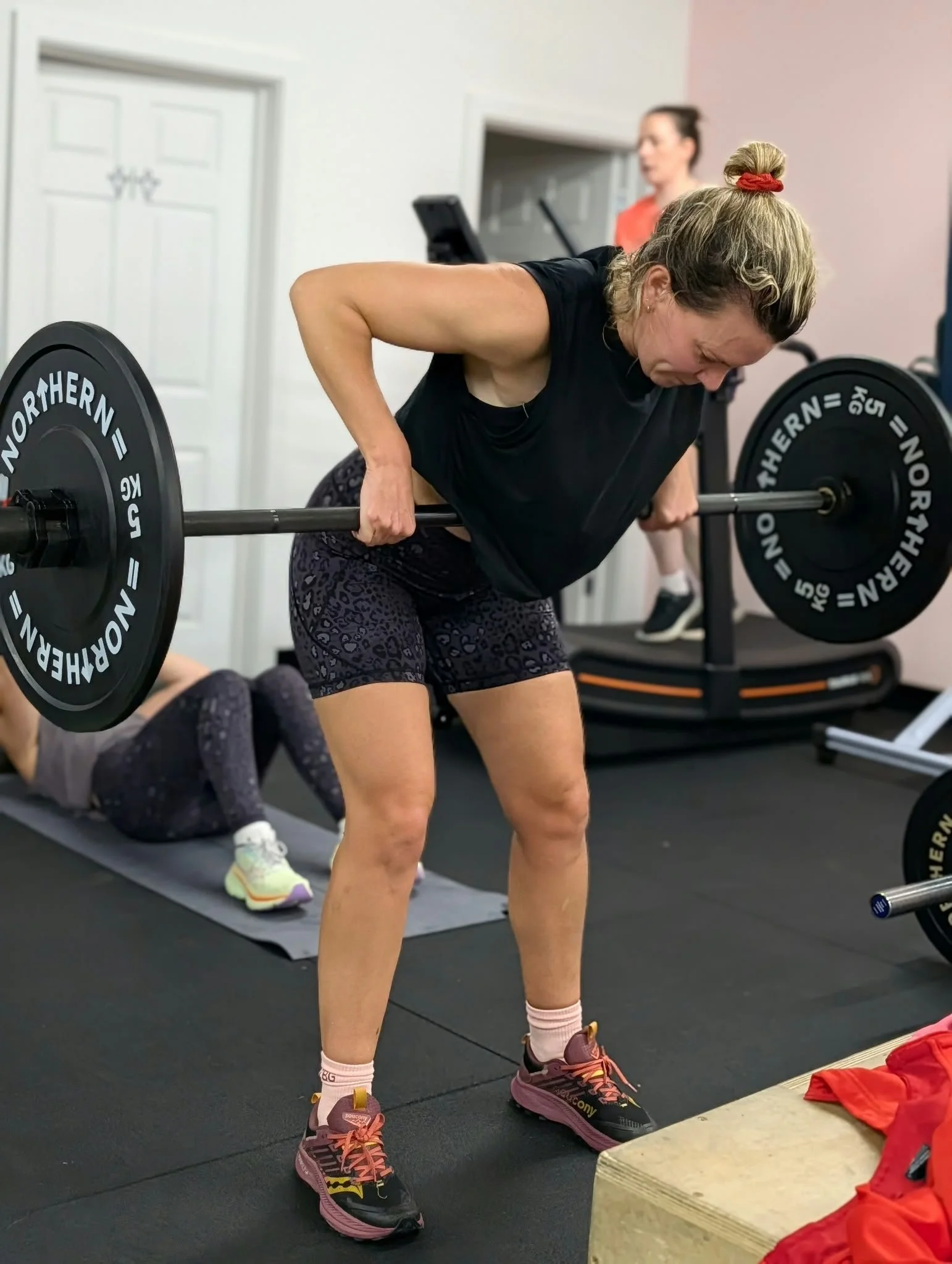 A woman lifting a barbell with weights in a gym, wearing workout clothes and sneakers, with a woman on a treadmill in the background.