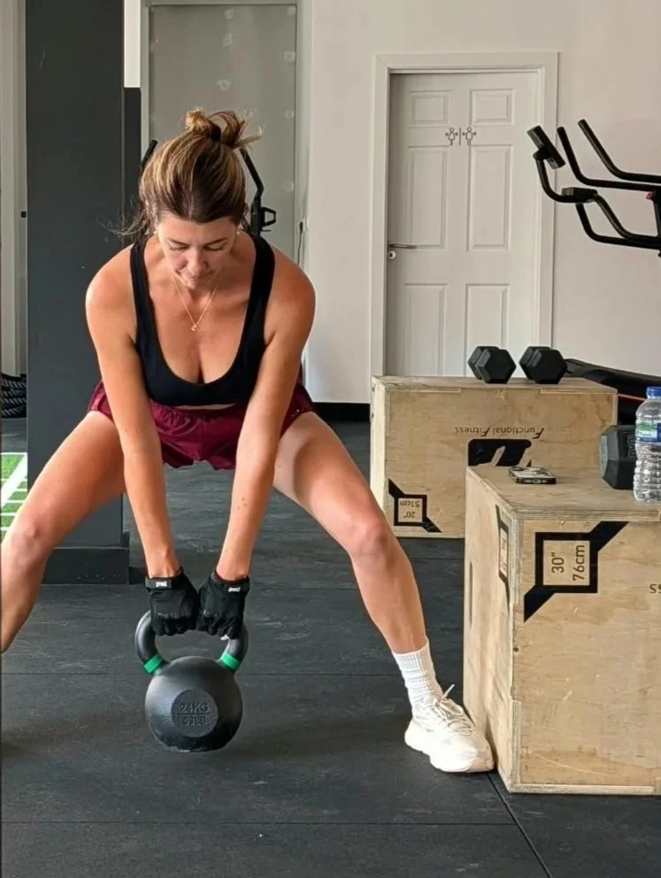 Woman performing kettlebell exercise in a gym, with weights and workout equipment around.