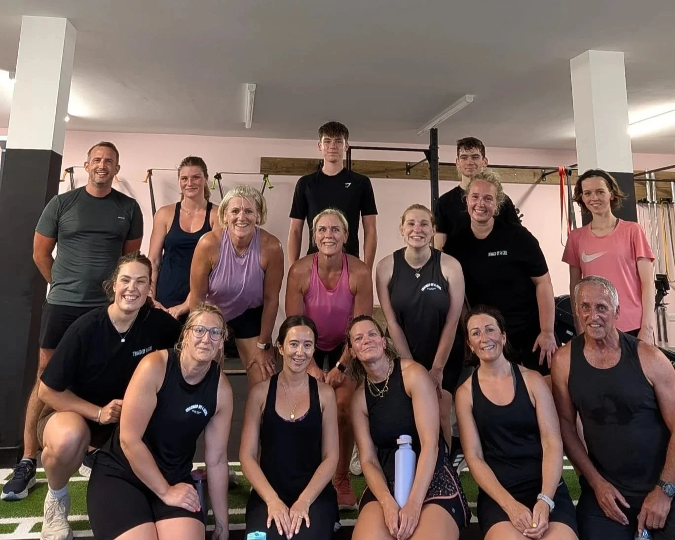 Group of diverse people in a gym posing for a photo, some kneeling and some standing, smiling, with workout equipment in the background.