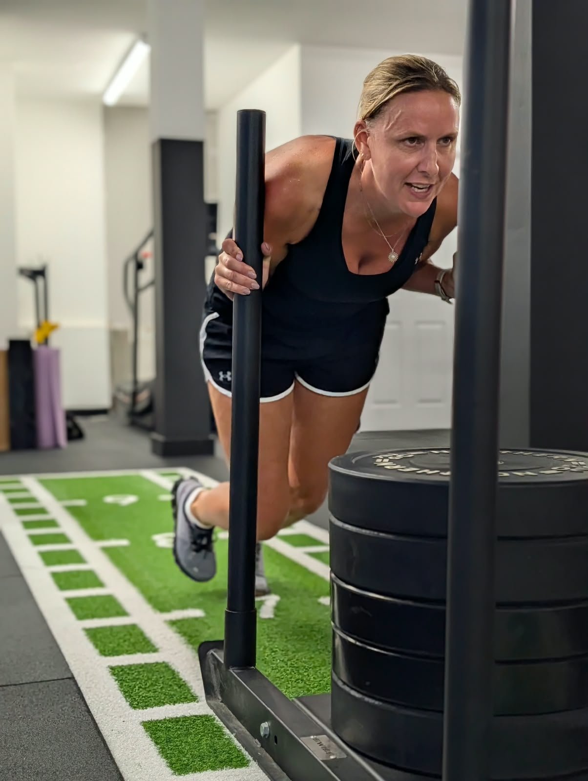 Woman doing a sled push exercise in a gym, pushing a sled with weights on a green and white workout mat.