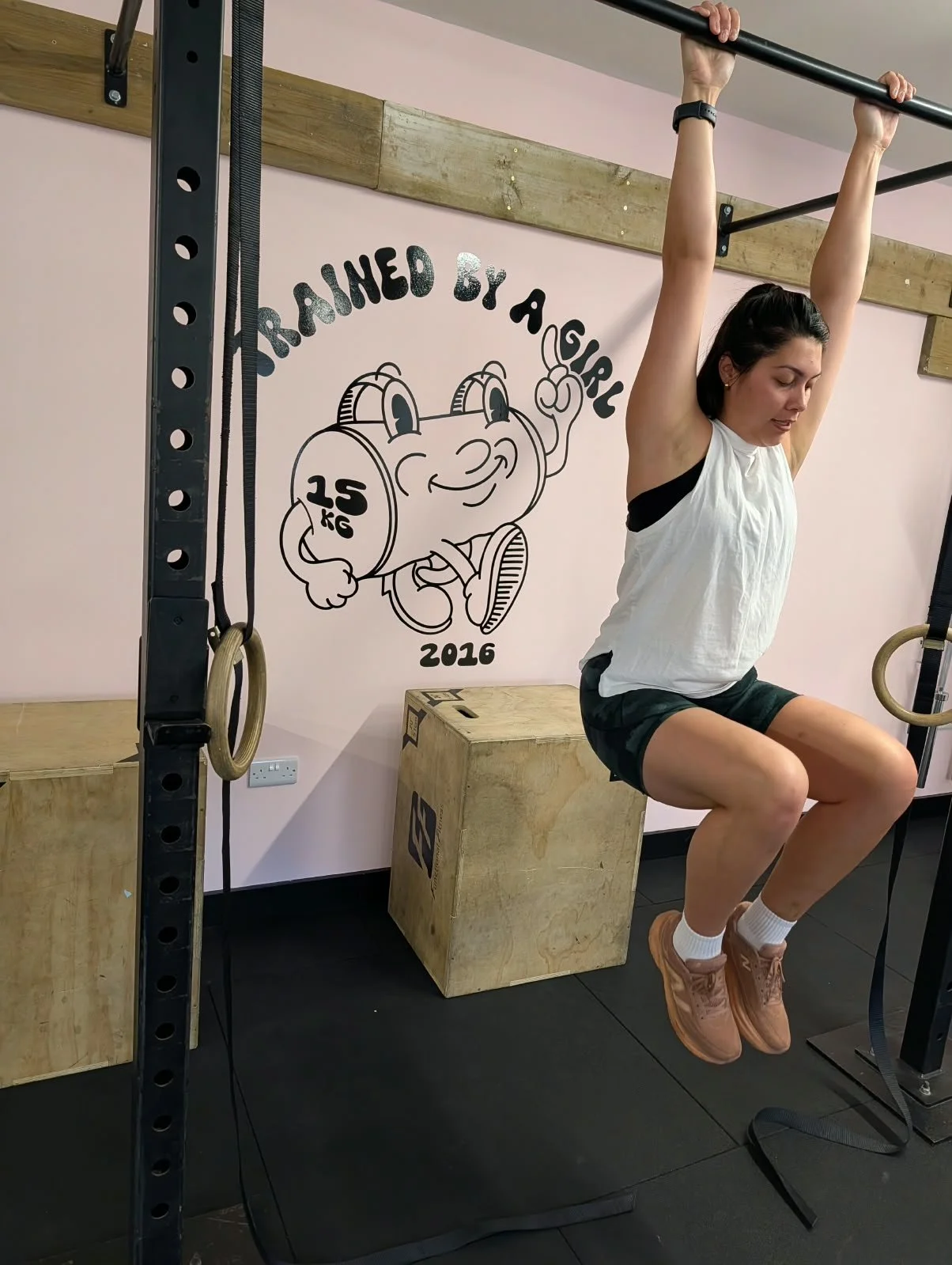 A woman performing an exercise on gymnastic rings hanging from a black metal frame, with wooden plyometric boxes nearby, in a gym with a wall illustration of a smiling cartoon weight with glasses and the text 'TRAINED BY ALEXU 2016'.