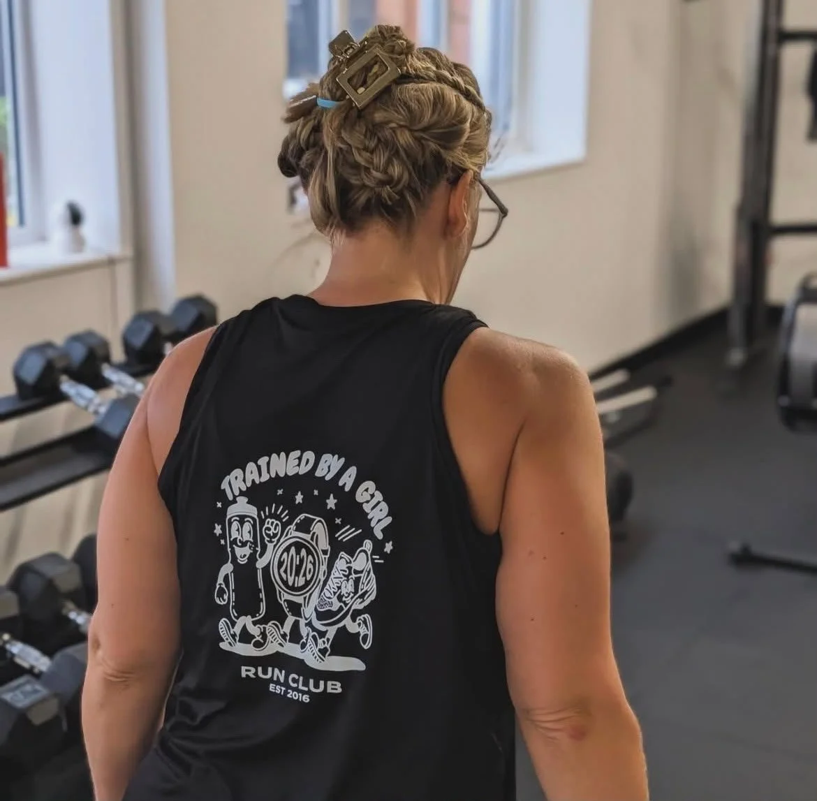 Back of a woman with blonde braided hair, wearing glasses and a sleeveless black shirt with a logo that reads 'Trained by a Girl' and 'Run Club.' She is in a gym with dumbbells on racks and gym equipment in the background.