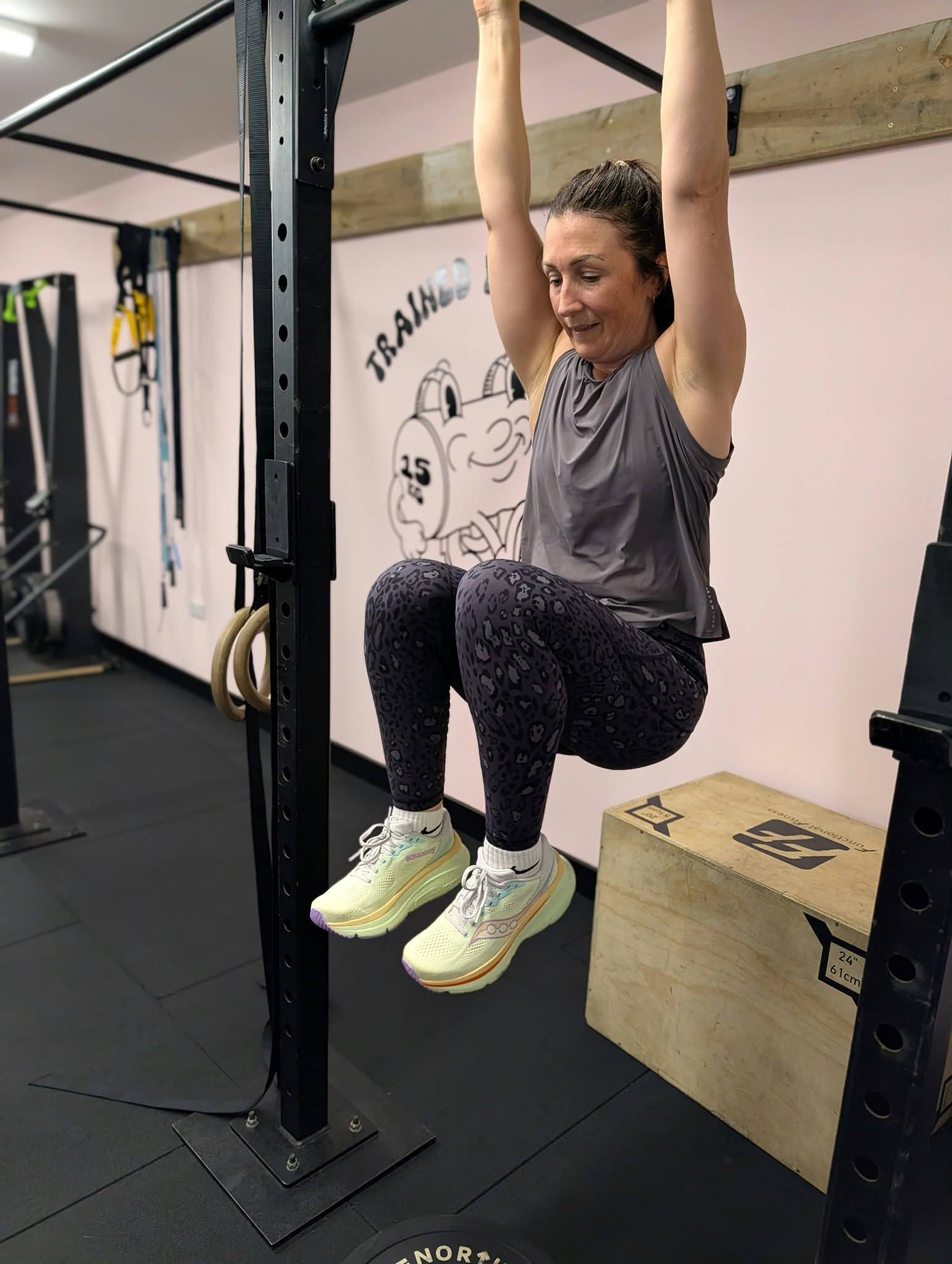 A woman in athletic wear, lifting her legs and holding onto a pull-up bar in a gym. She is wearing gray tank top, leopard-print leggings, and white sneakers, with gym equipment and a wall with gym-related drawings in the background.