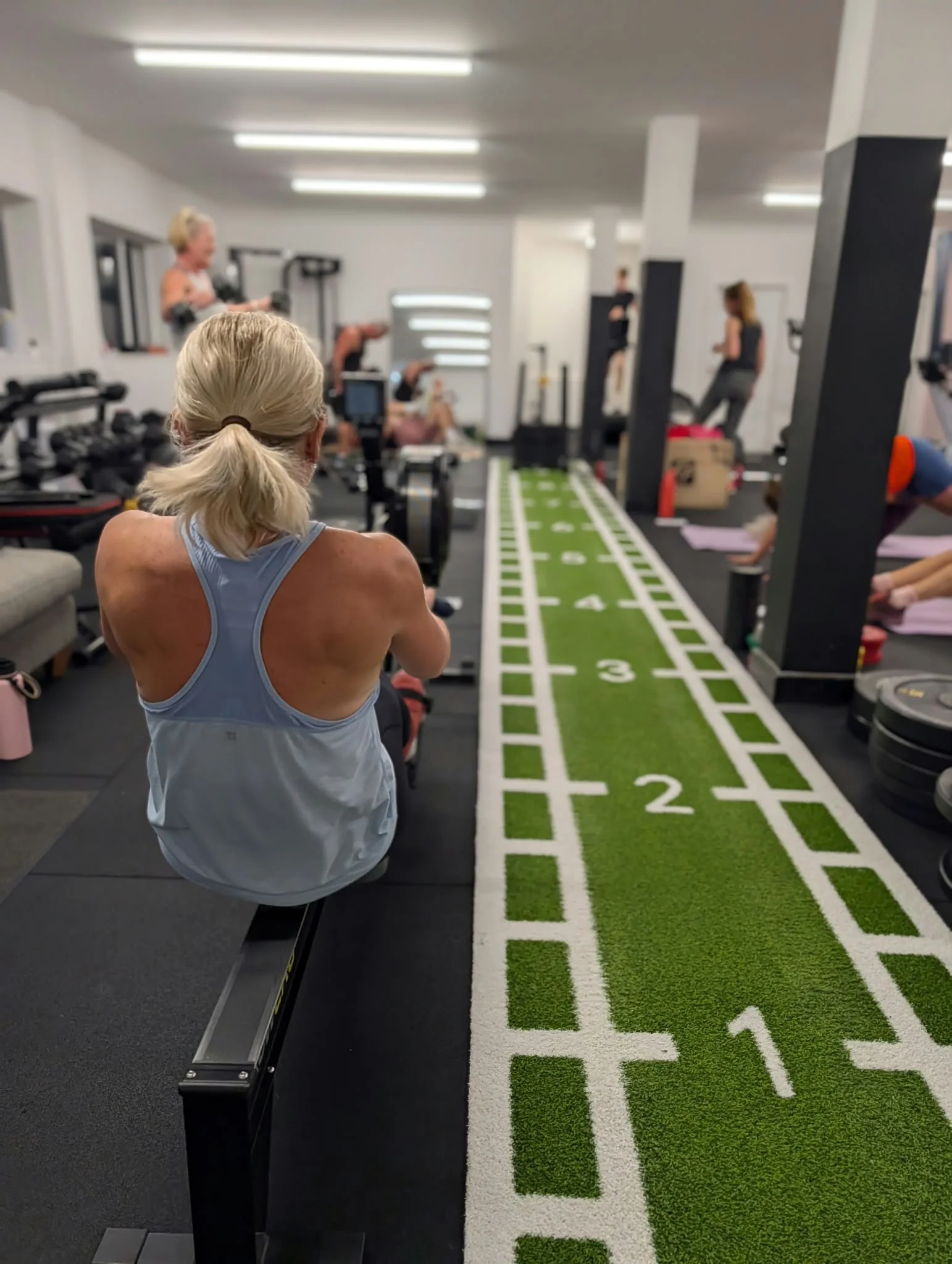 People participating in a fitness class in a gym, sitting on workout benches and stretching on a green and white strip-style training mat.