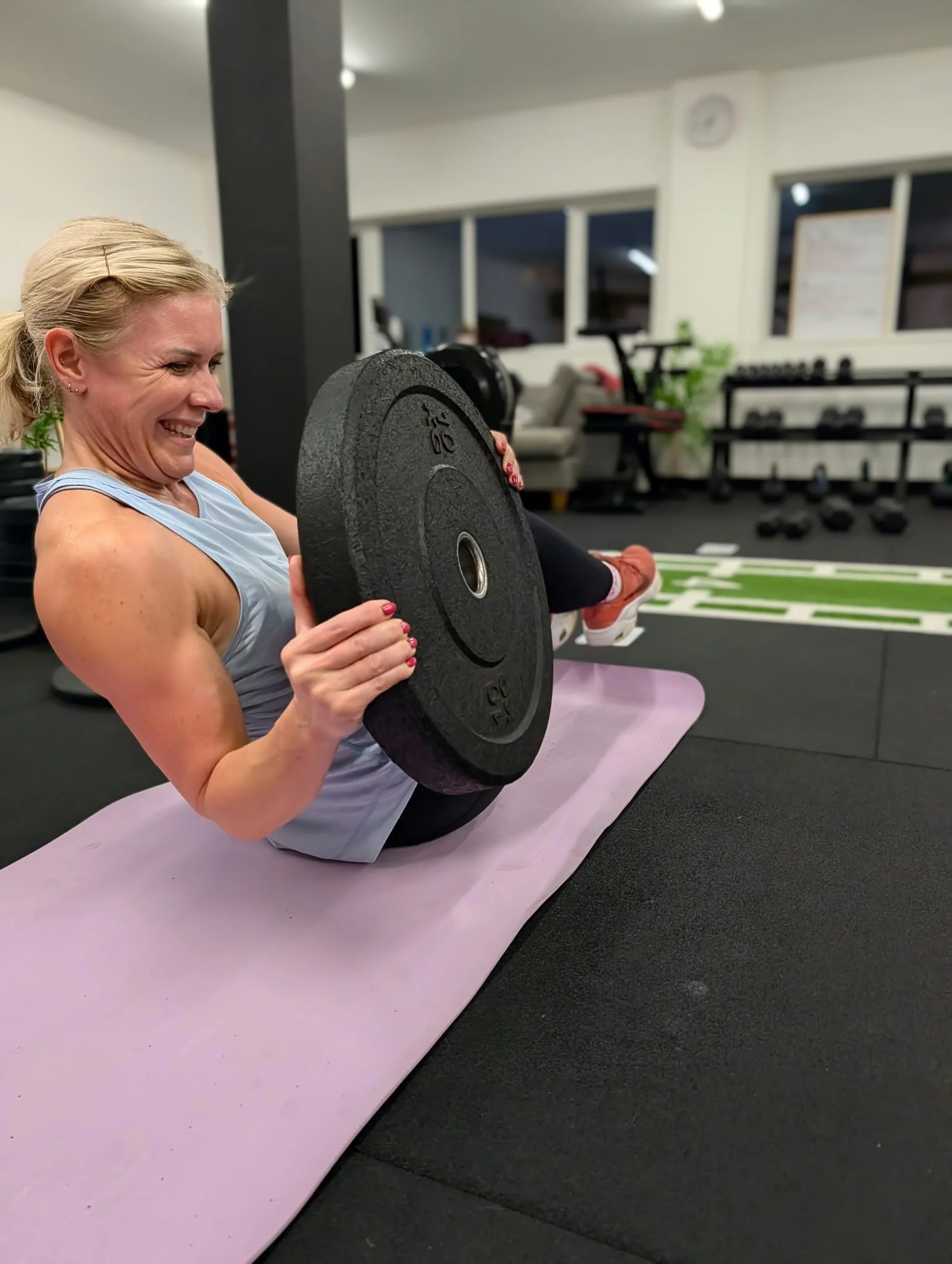 A woman in workout clothes doing a leg curl exercise on a workout mat in a gym.