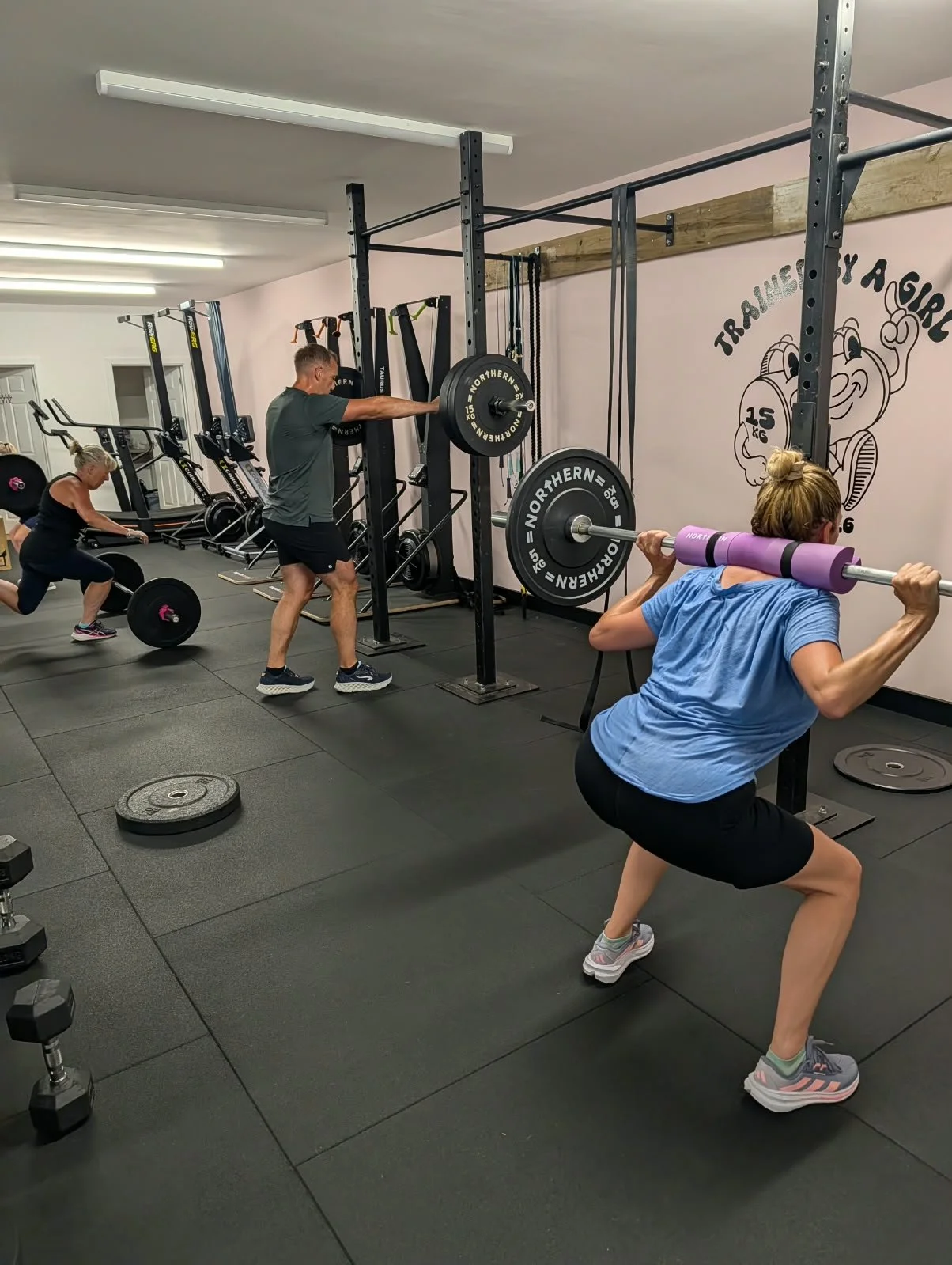 People working out in a gym, lifting weights and doing squats. The gym has black flooring, weight plates, and wall decor with the phrase 'Trainery A Gym.'