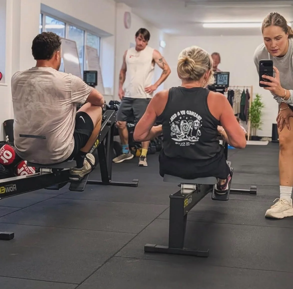 People working out in a gym, with one woman seated on a rowing machine, a woman standing next to her taking a selfie, and other individuals in the background.