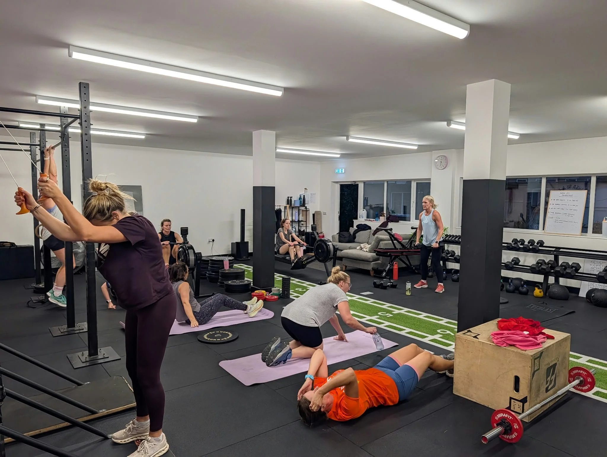 People participating in a fitness class in a gym, performing various exercises including stretching and rowing, with gym equipment and weights visible in the background.