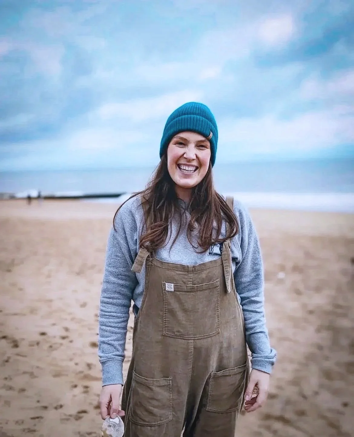 Smiling woman wearing a blue beanie, gray hoodie, and brown overalls standing on a sandy beach with ocean and cloudy sky in the background.
