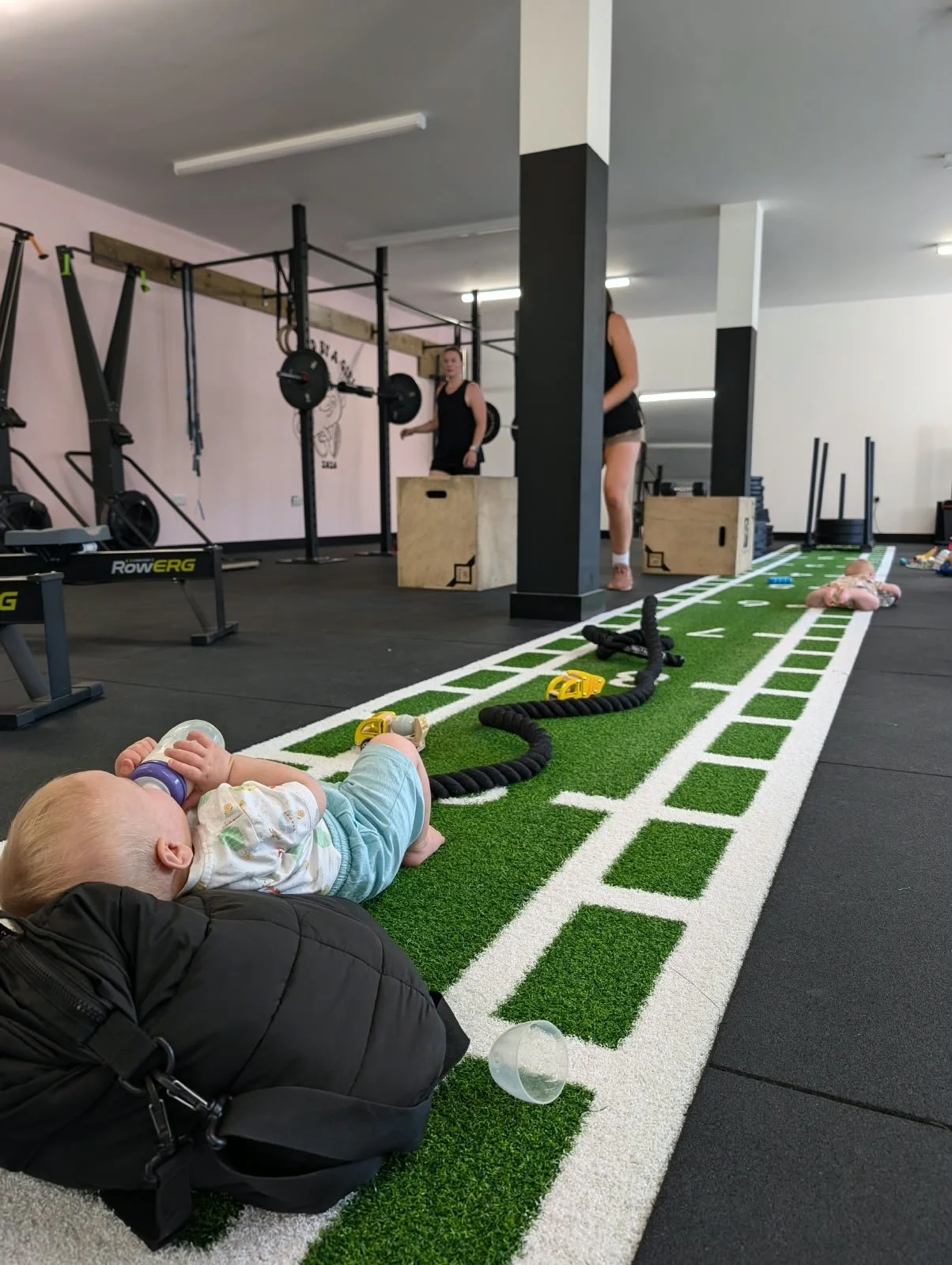 Child lying on a green turf track at a gym, with gym equipment and two women working out in the background.