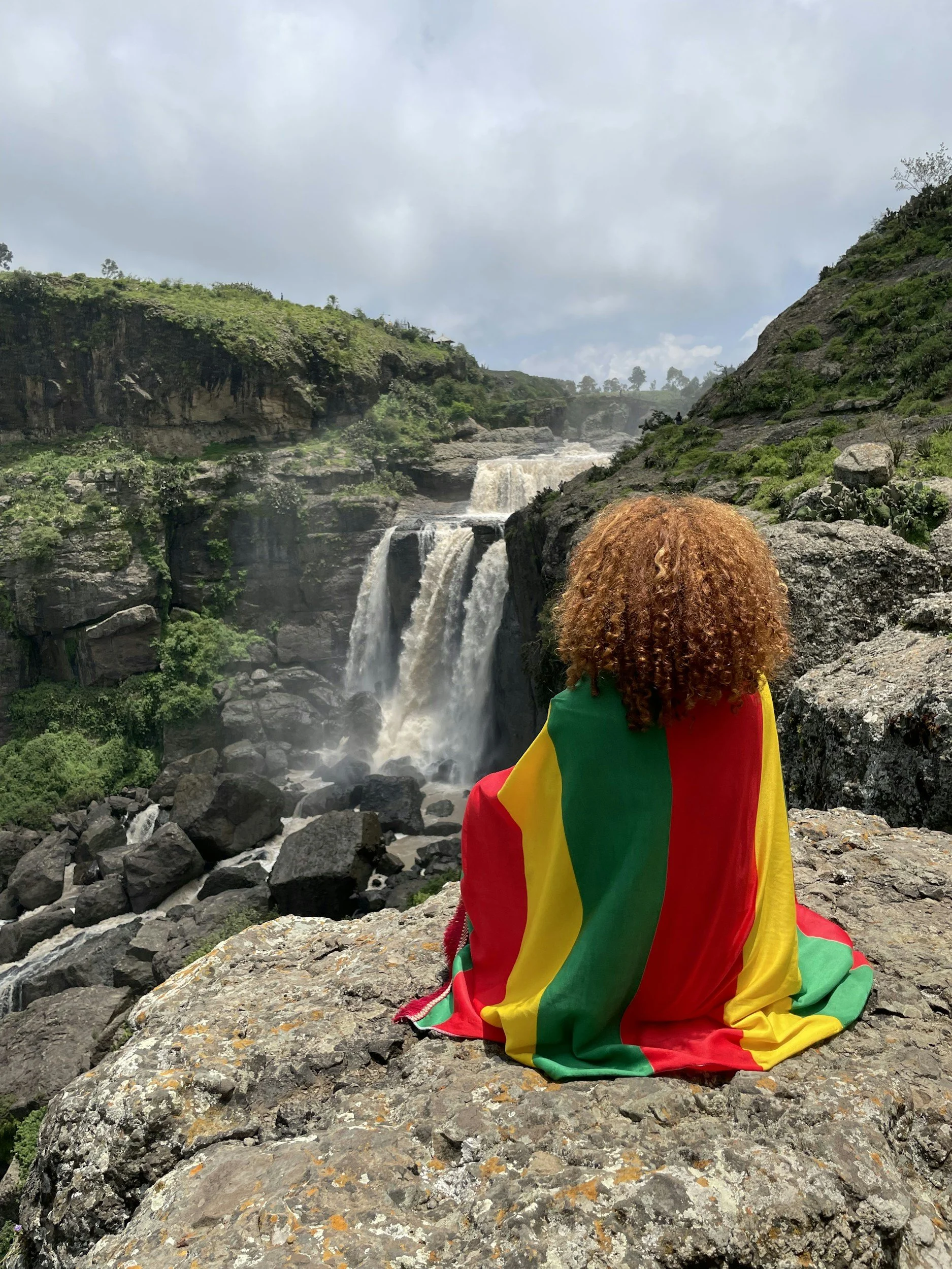 Person with curly hair draped in a flag sitting on rocky terrain in front of a waterfall with lush greenery and cloudy sky.