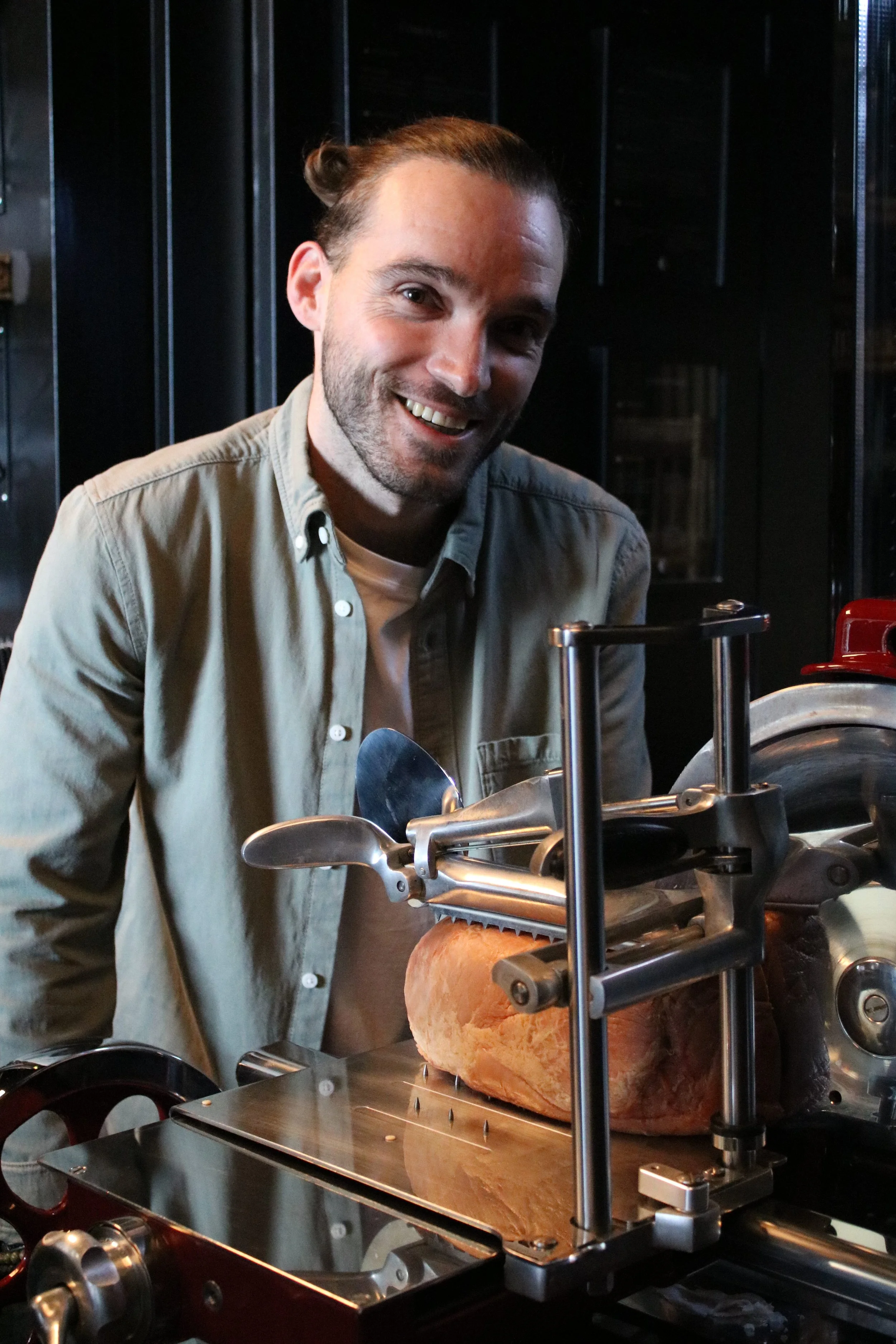 Homme avec barbe souriant, travaillant avec une machine à trancher du jambon ou du fromage dans une cuisine de G.APERO ANDREZIEUX