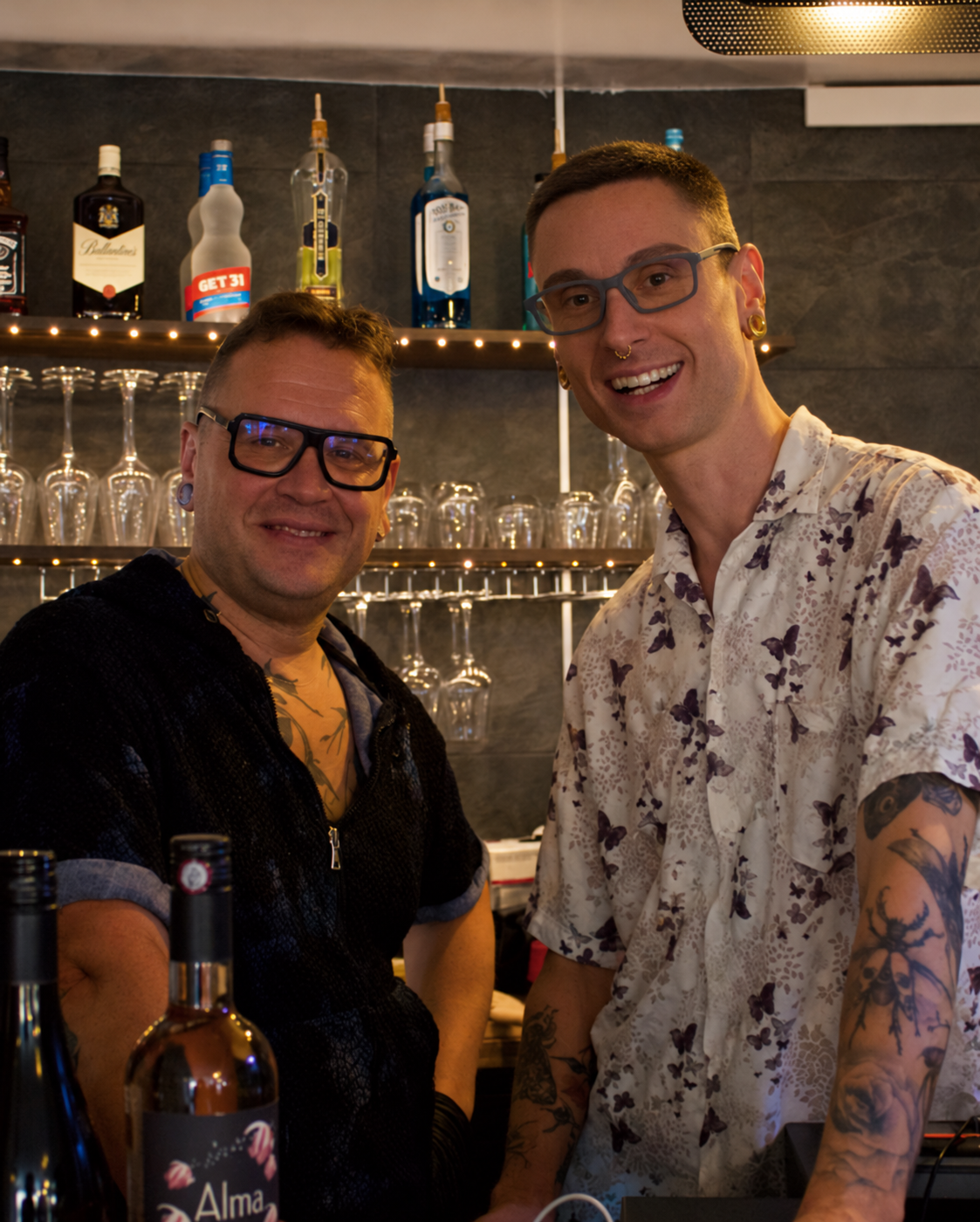 Deux hommes souriants posent dans un bar, avec des bouteilles et des verres derrière eux à G.APERO HYÈRES