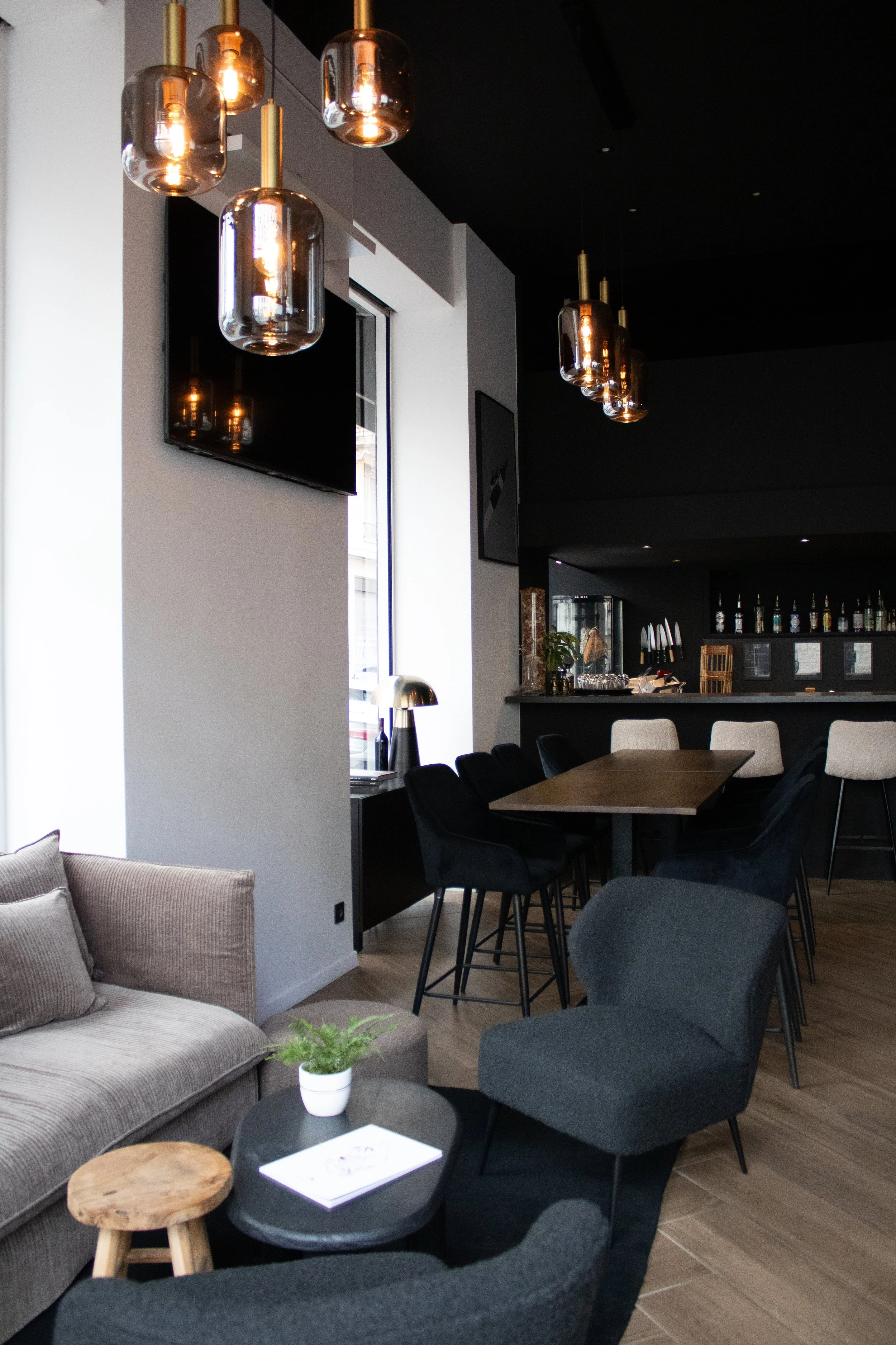 Intérieur de G.APERO LYON, un restaurant à Lyon 6eme avec des lampes suspendues en verre, un mur avec une télévision, des tables et fauteuils modernes, un comptoir en arrière-plan, et un décor sobre et élégant.