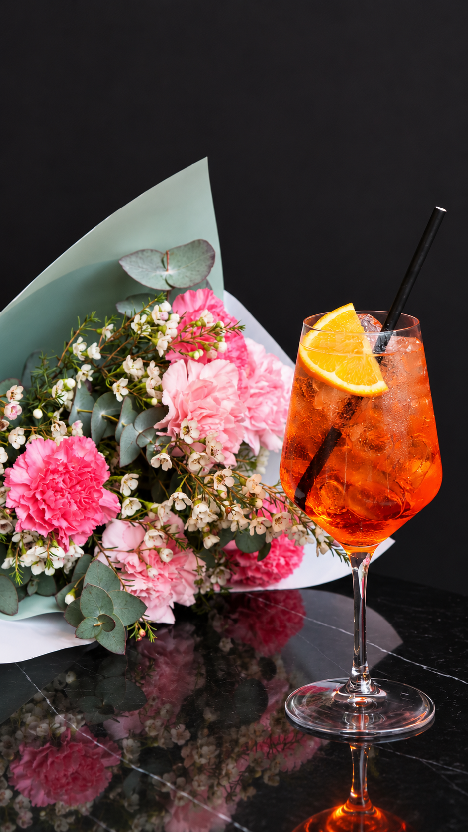 Un verre de cocktail avec des glaçons et une tranche d'orange, à côté d'un bouquet de fleurs roses et blanches, sur une surface en marbre noir.