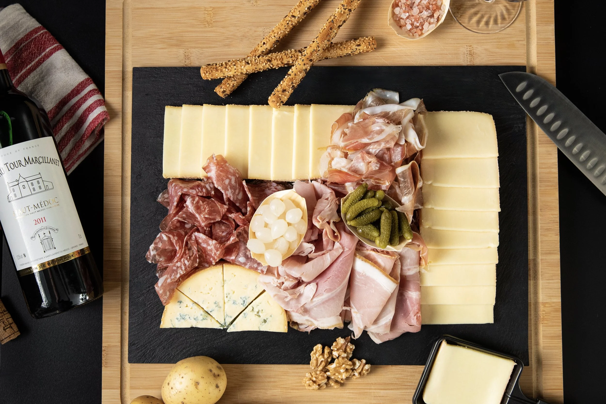 Planche Raclette : fromages, charcuterie, cornichon, noix, pommes de terre et vin, vue du dessus.