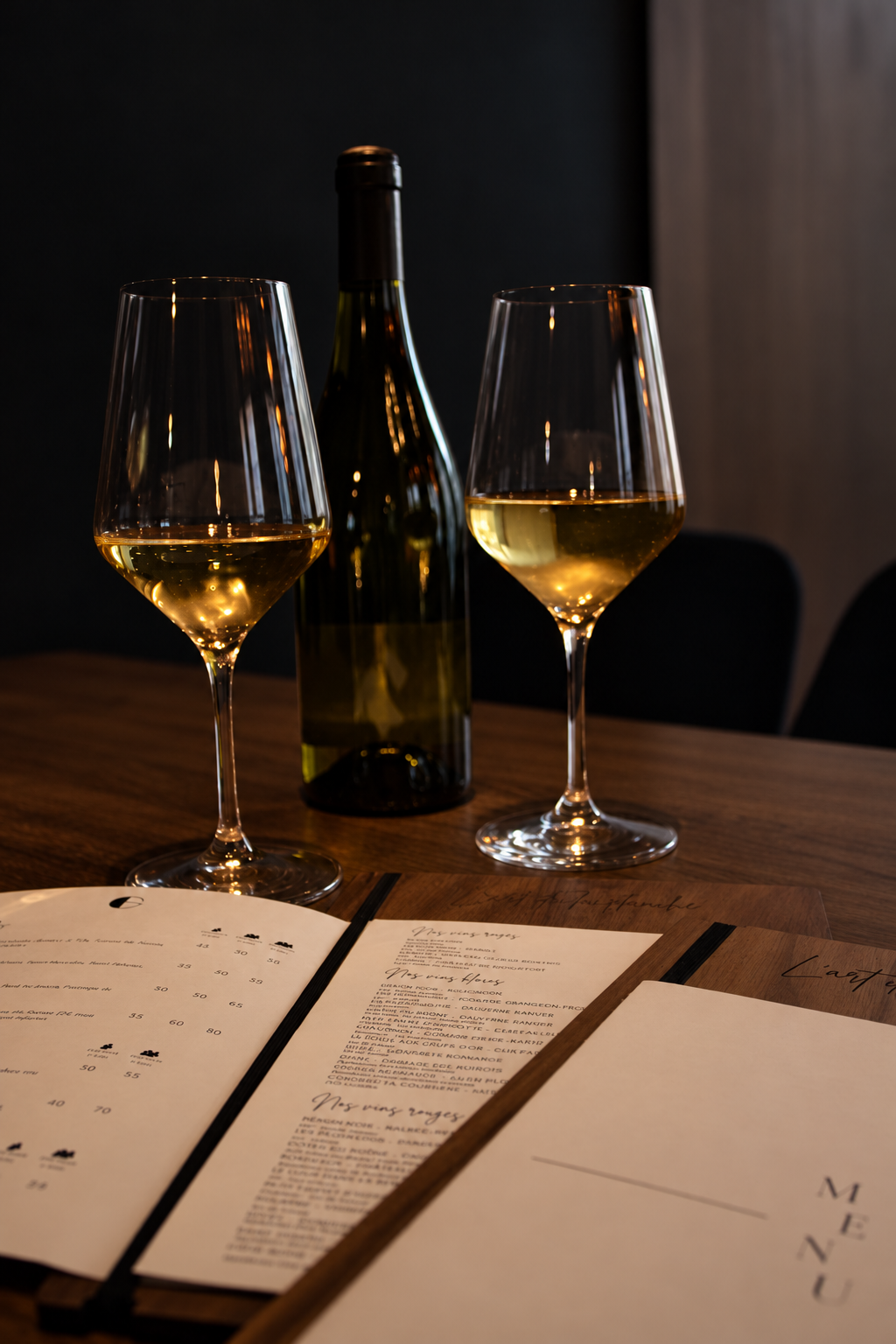 Deux verres de vin blanc sur une table en bois, avec une bouteille de vin en arrière-plan et des menus déroulés devant. L'ambiance est tamisée, créant une atmosphère chaleureuse.
