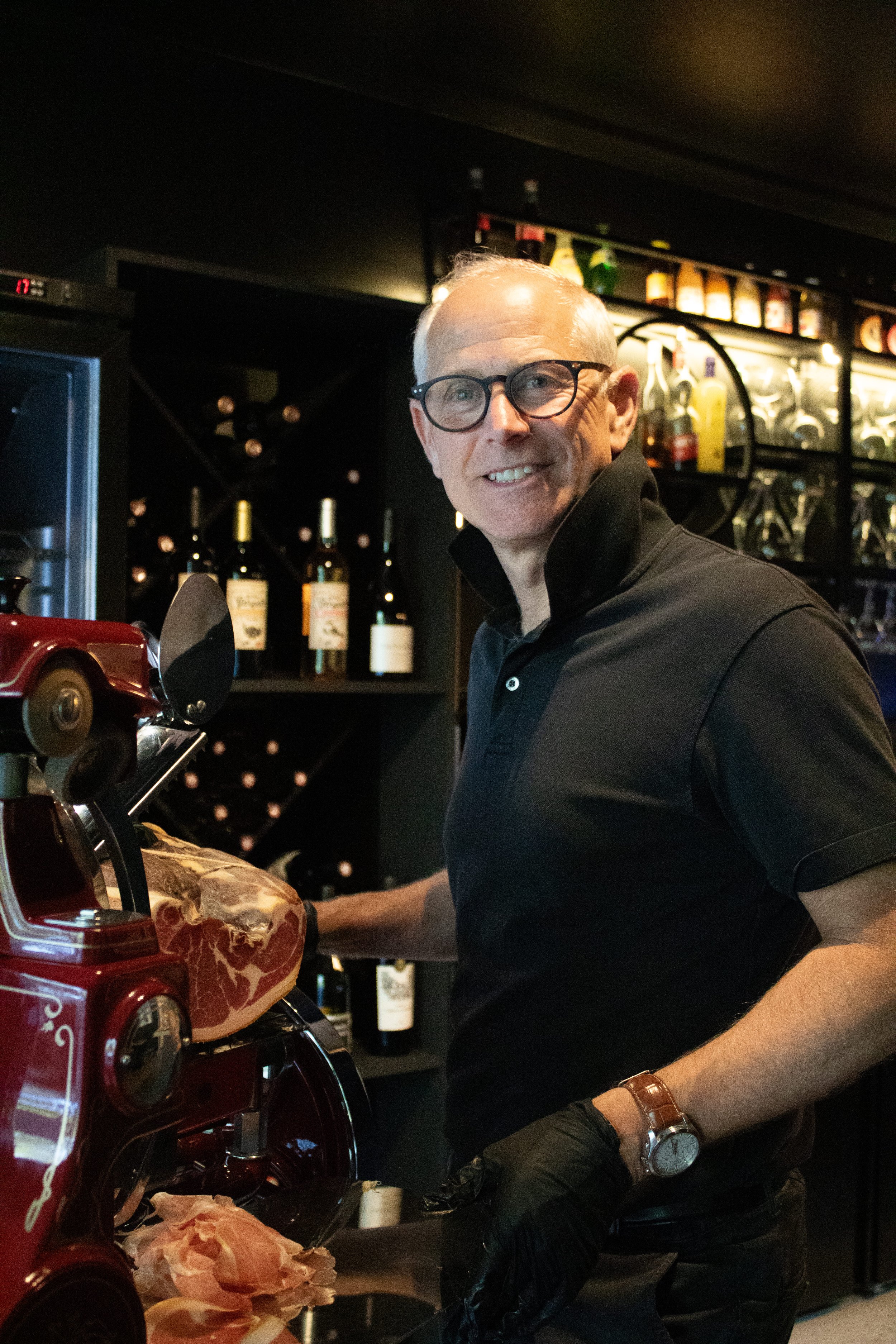 Un homme souriant portant des lunettes noires, un costume noir, en train de préparer de la viande derrière un comptoir dans un bar ou un restaurant, avec des bouteilles d'alcool en arrière-plan à G.APERO LA CELLE SAINT CLOUD