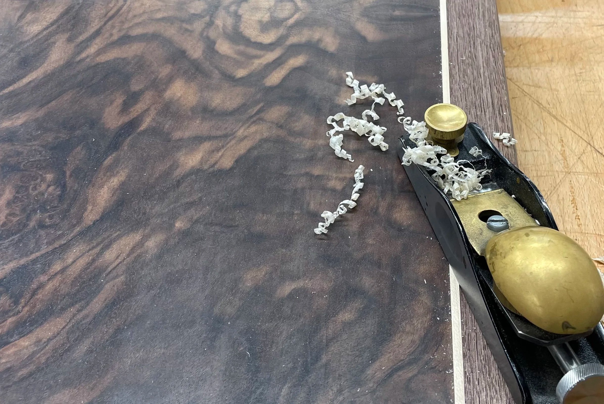 Close-up of a block plane with curled wood shavings being used to finish a table top made from walnut, walnut burr veneer and maple inlay .