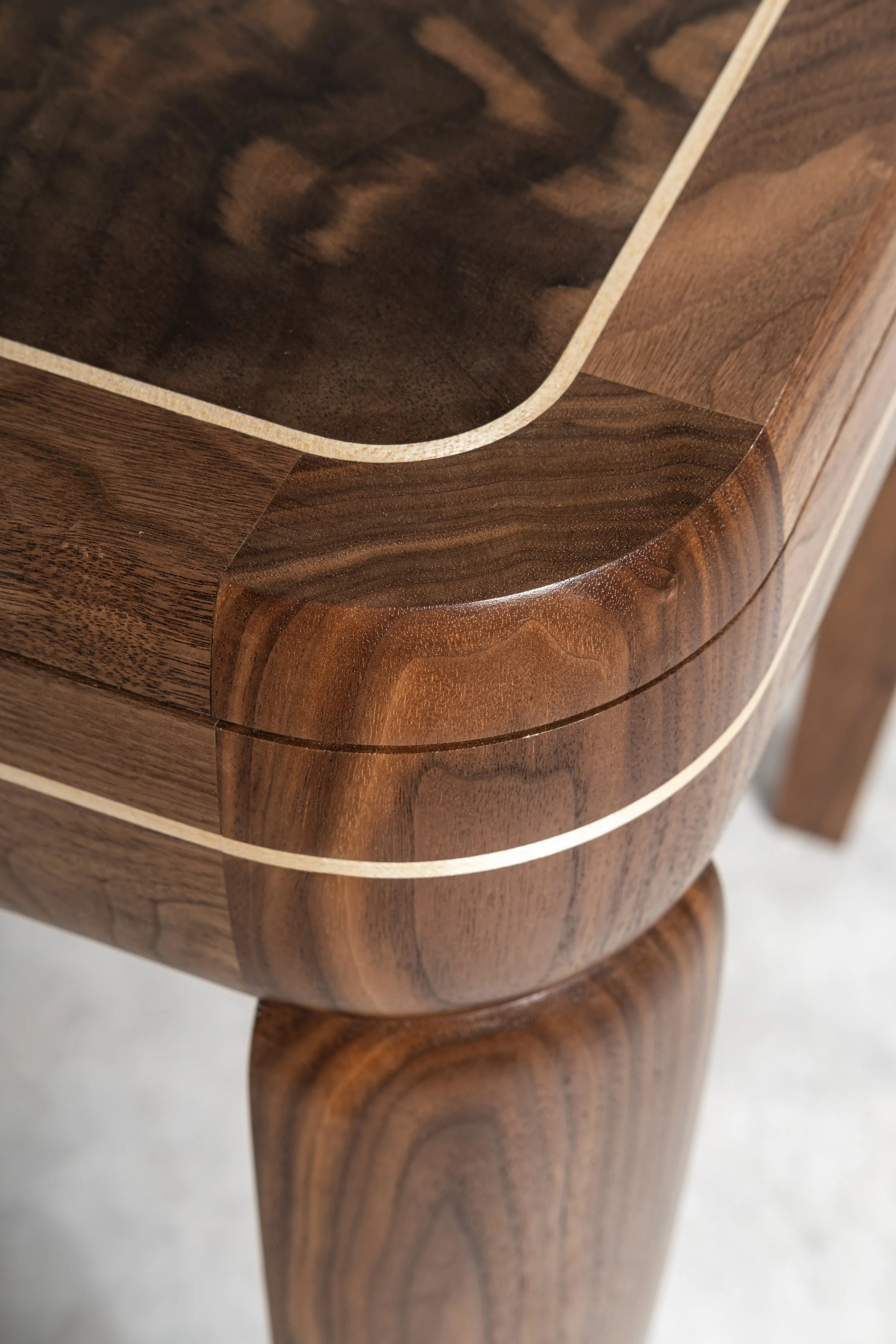 Close-up of the corner of the Malone Desk showing a single quarter-turned walnut leg with beautiful grain pattern, joining walnut aprons and walnut burr veneer top and walnut inlay detail
