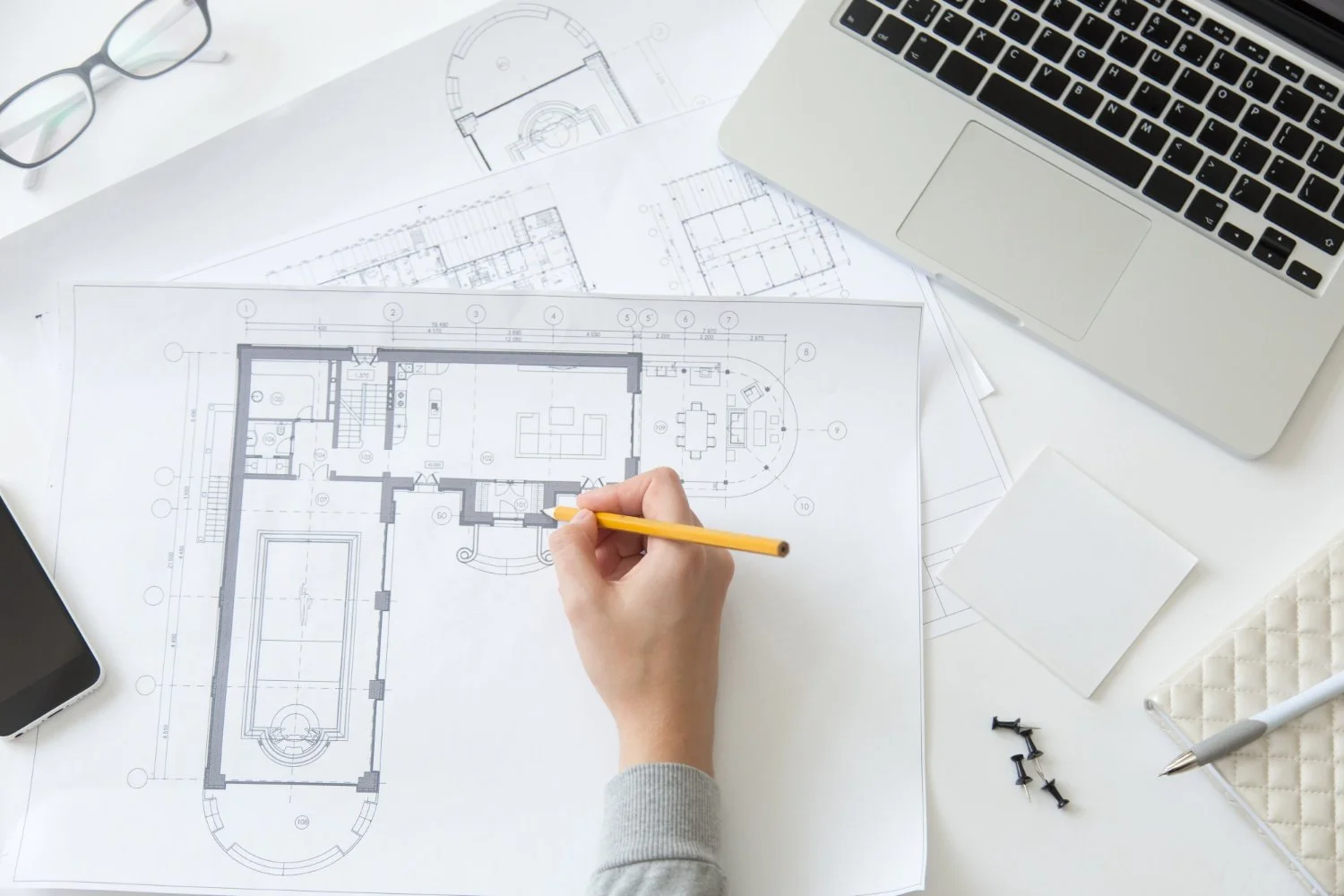 Hand drawing a detailed floor plan with architectural blueprints and a laptop for furniture placement and senior downsizing space planning.