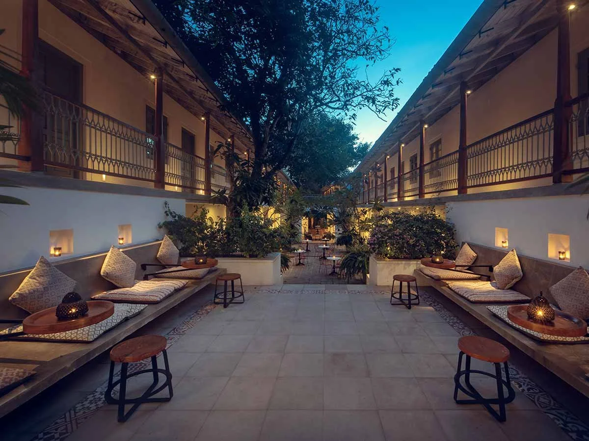 Elegant courtyard lounge with cushioned benches, stools, plants, and warm lantern lighting at dusk
