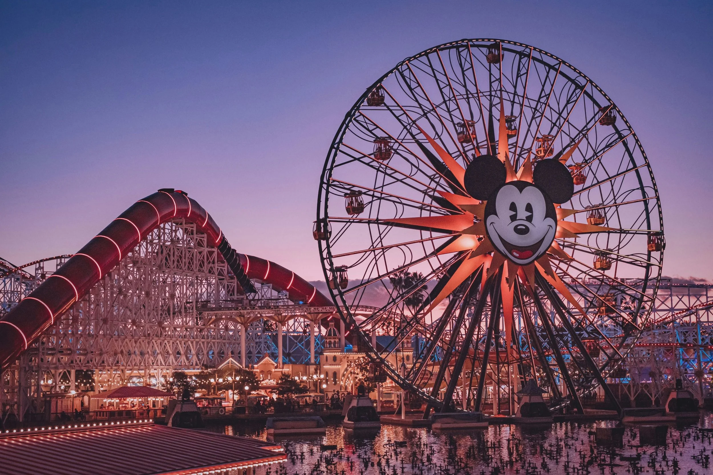 A Disney California Adventure amusement park Ferris wheel named Mickey's Fun Wheel with Mickey Mouse's face at the center, illuminated with orange lights during dusk, along with a roller coaster with red tracks in the background.