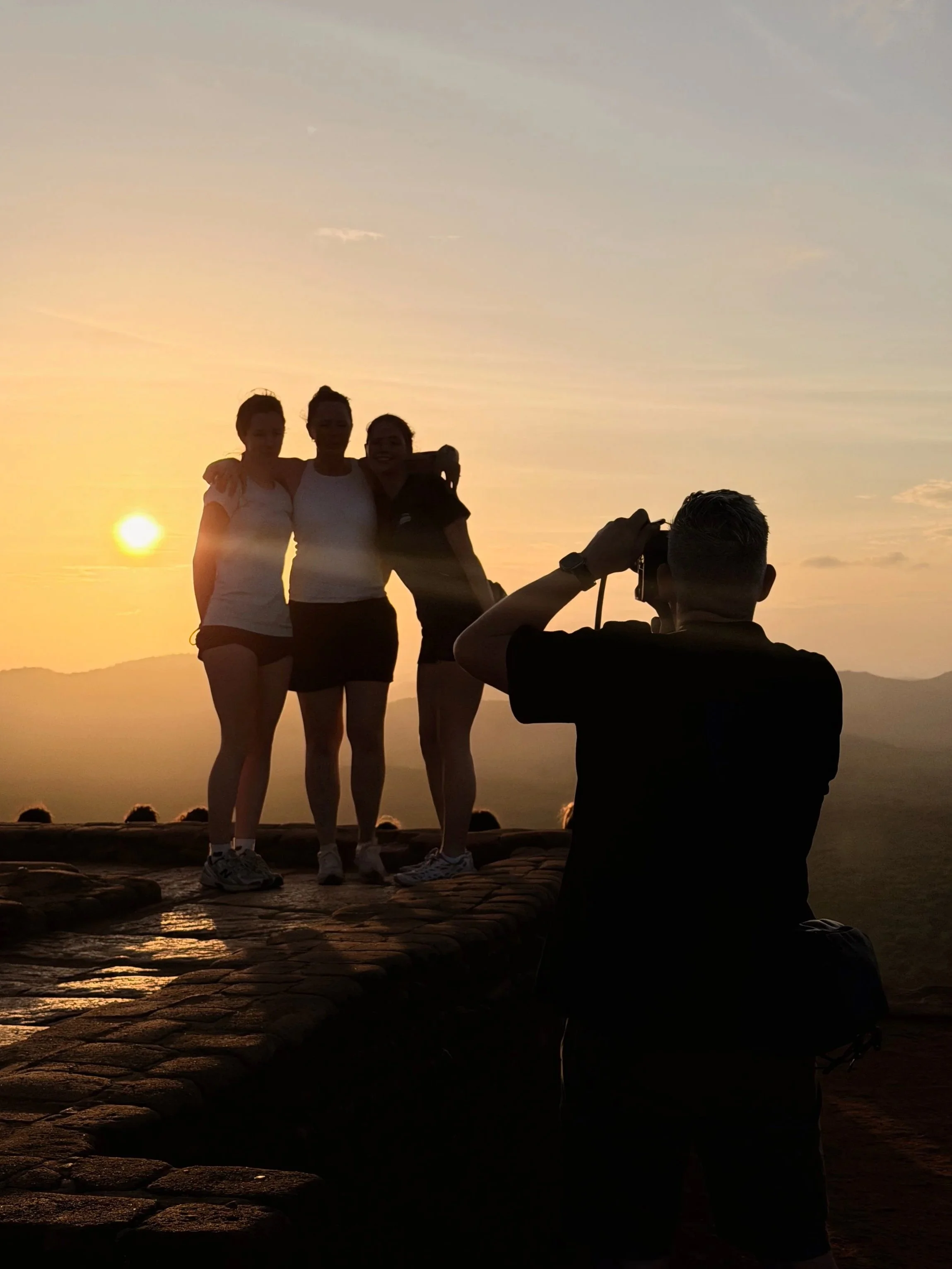 Four people are standing together at sunset, with a host taking their photo. The sunset and mountains are visible in the background.