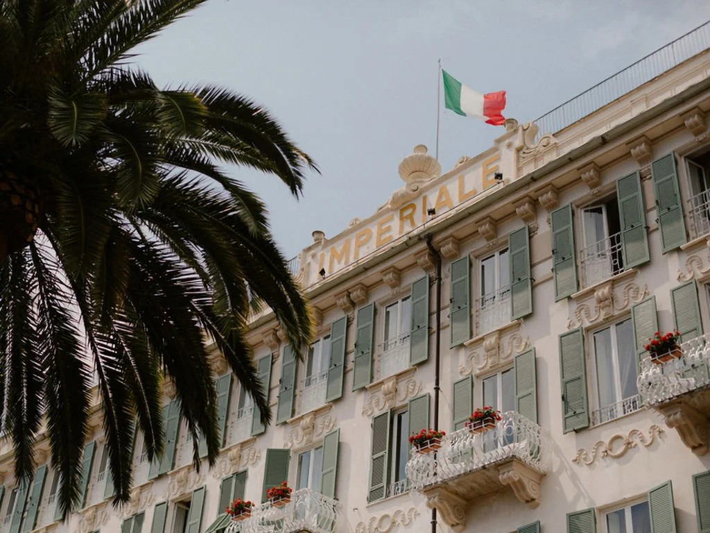 The front of a historic building with green-shuttered windows, small balconies with red flowers, and an Italian flag flying on top. The sign on the building reads 'Imperiale'.