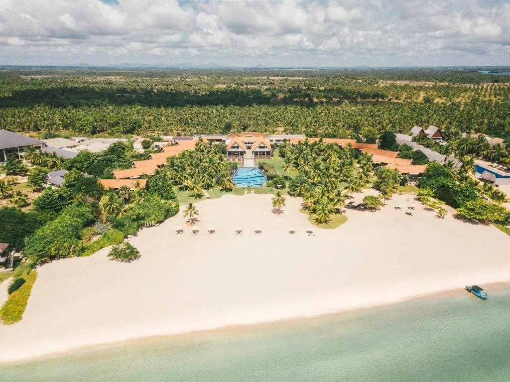 Aerial view of a tropical resort with sandy beach, palm trees, swimming pool, and ocean, surrounded by lush greenery and dense forest.