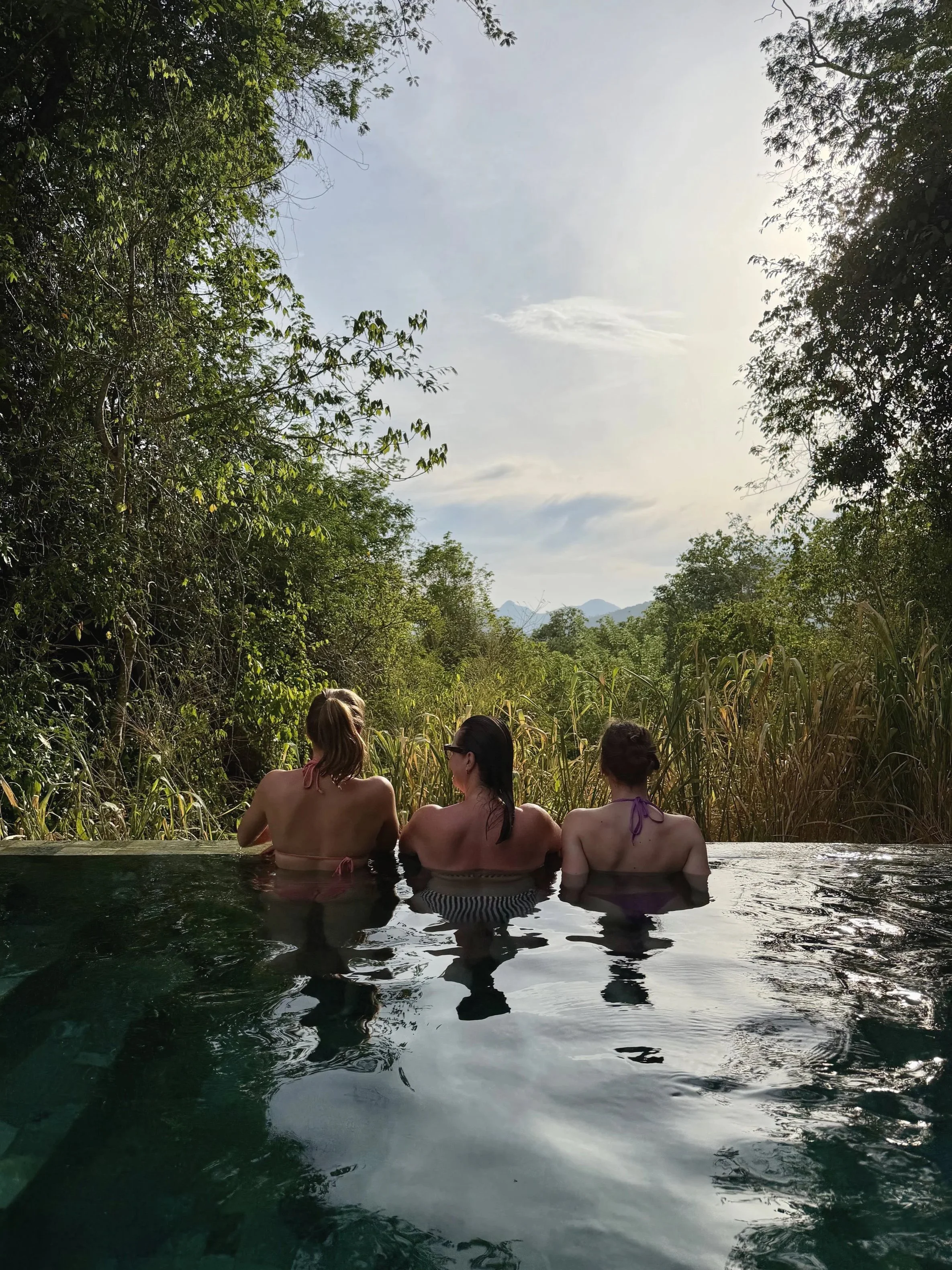 Three women sit in a hot spring pool surrounded by lush greenery, with a mountain in the background, during daytime.