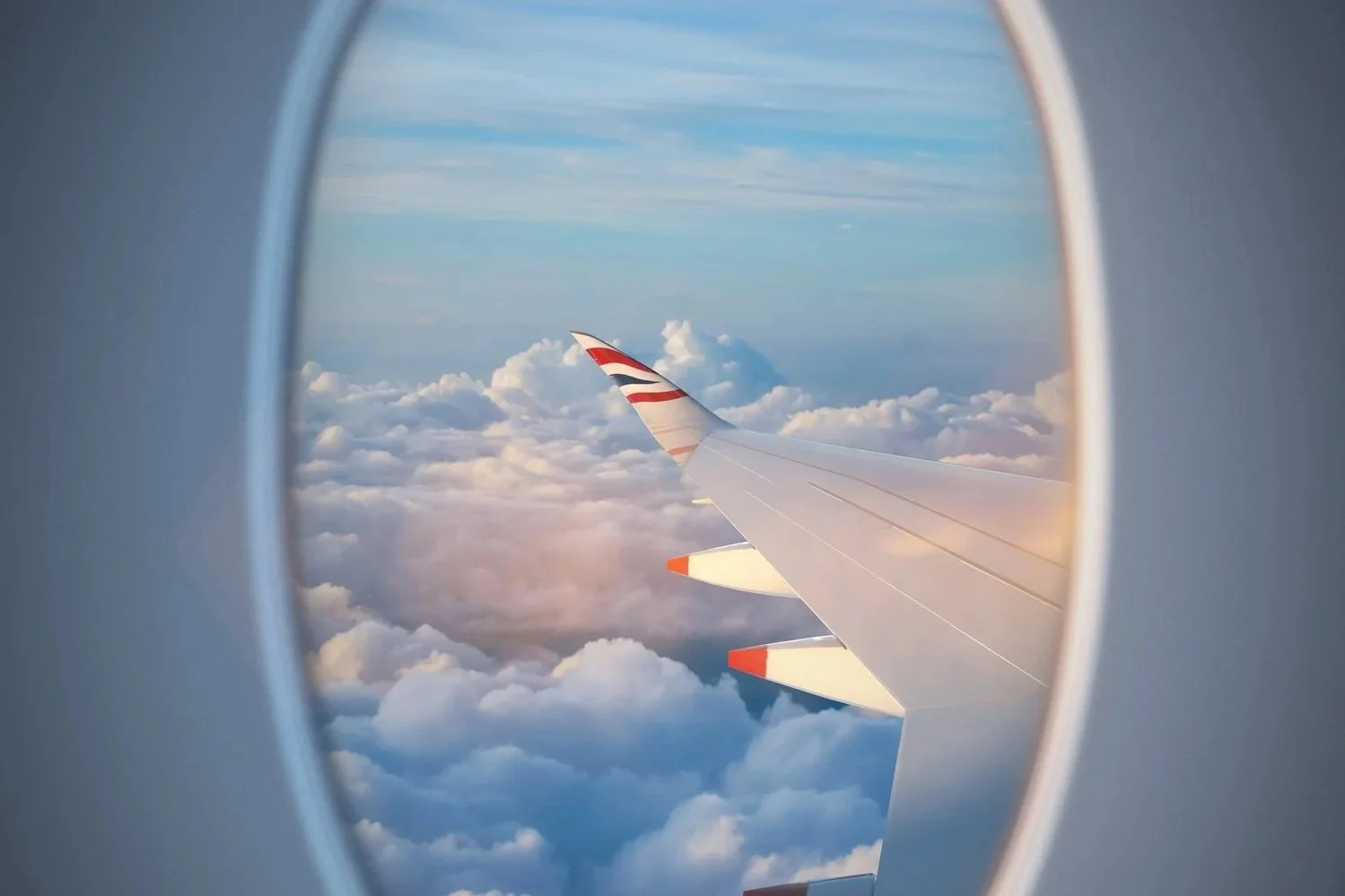 View of an airplane wing through a window, with clouds and blue sky in the background.