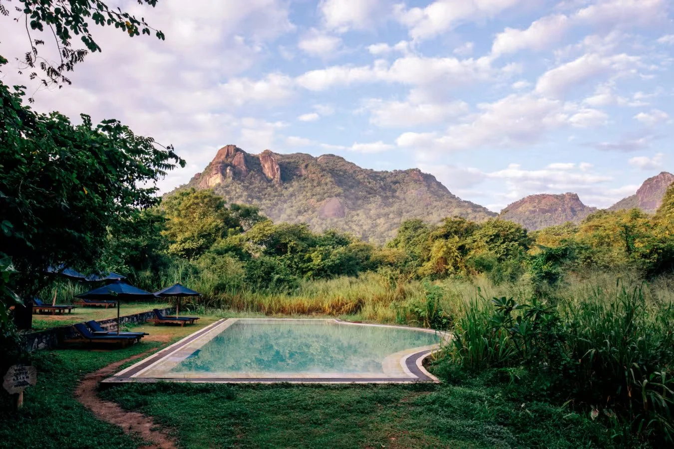 A swimming pool surrounded by greenery and lounge chairs with umbrellas, set against a backdrop of mountains and blue sky with clouds.