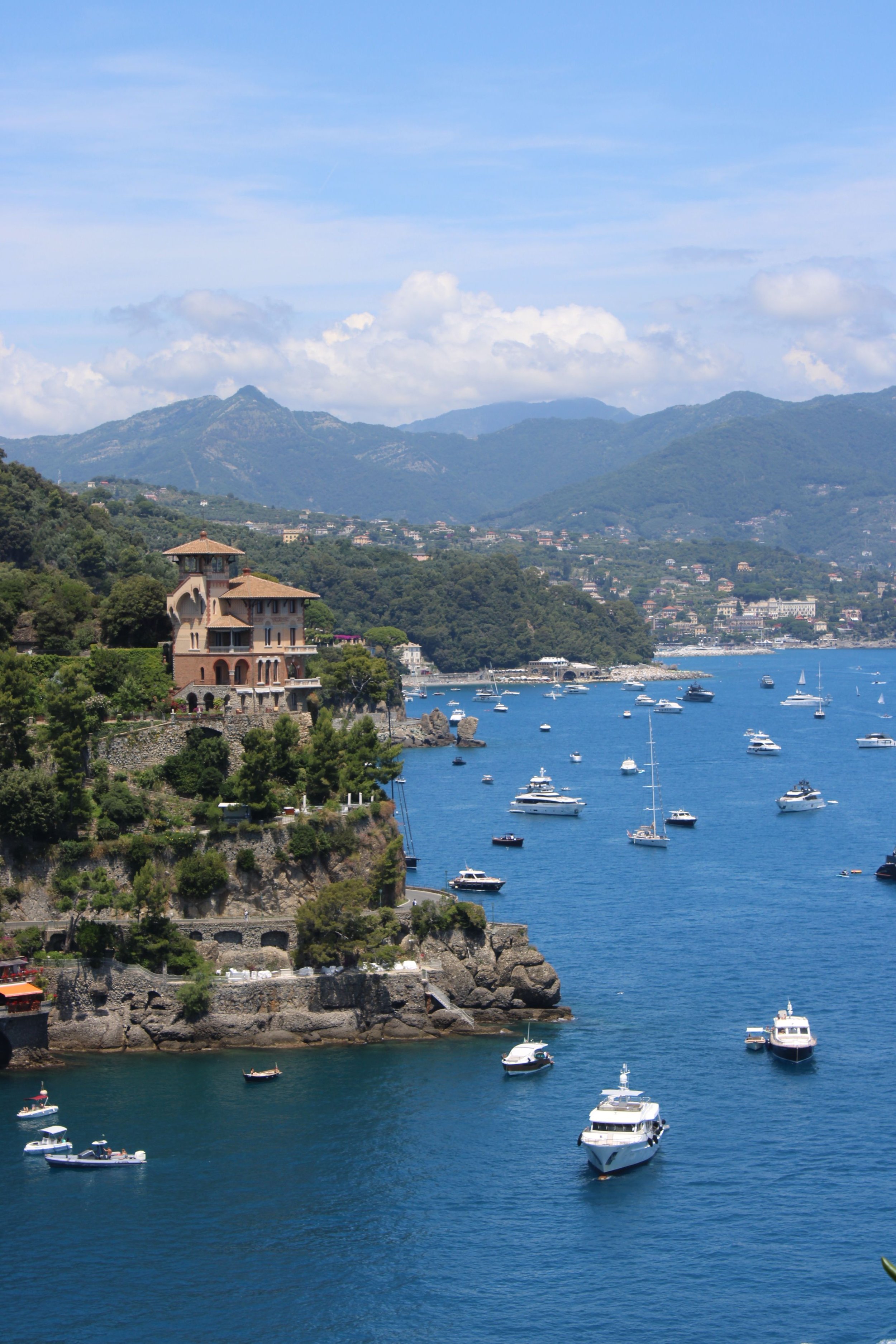 Scenic view of a hillside with a Mediterranean-style house overlooking a bay filled with numerous boats and yachts, green mountains in the background, and a partly cloudy sky.