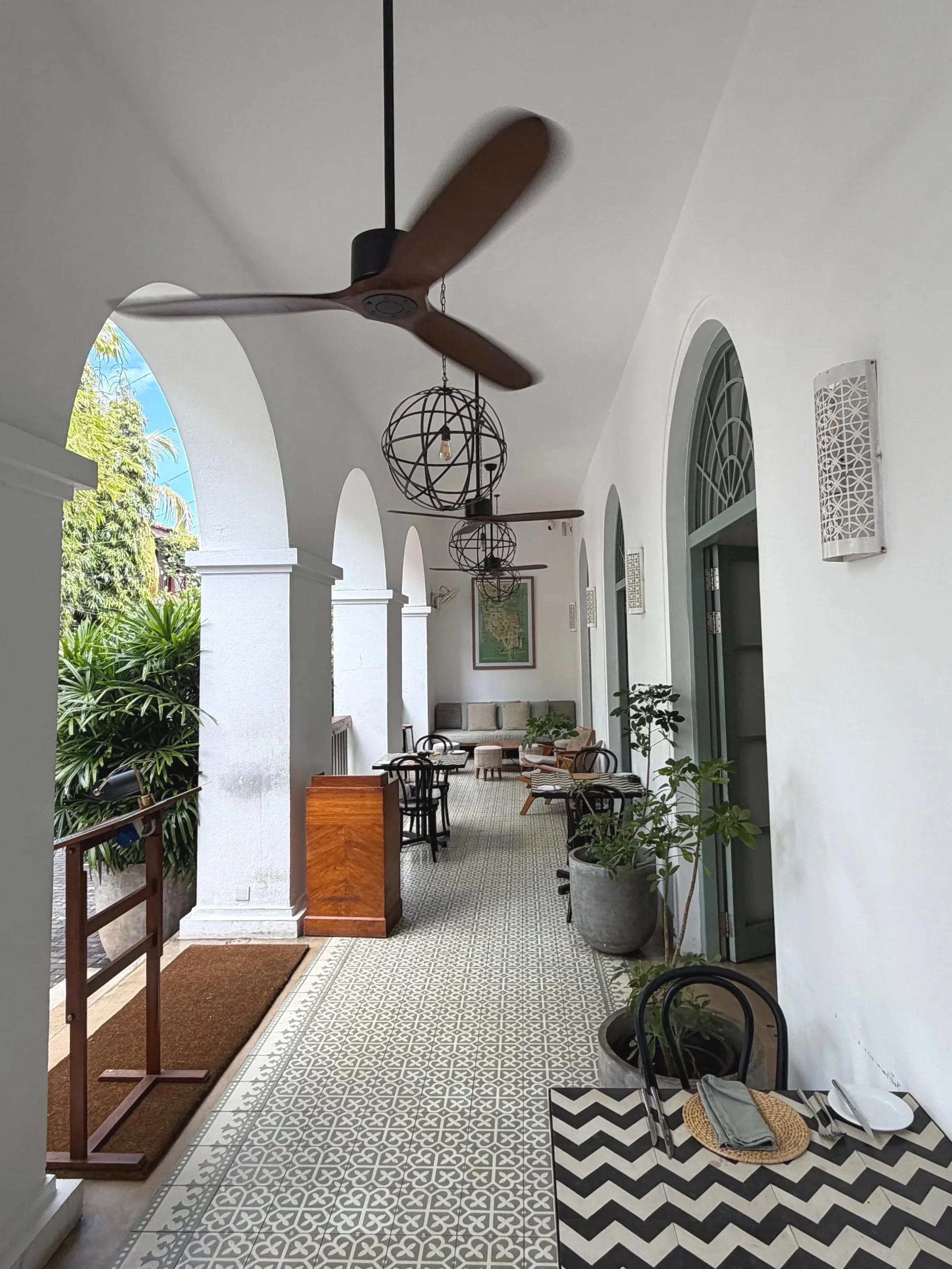 Covered outdoor patio with white arches, ceiling fans, geometric patterned tiles, potted plants, seating area, and a welcome sign.