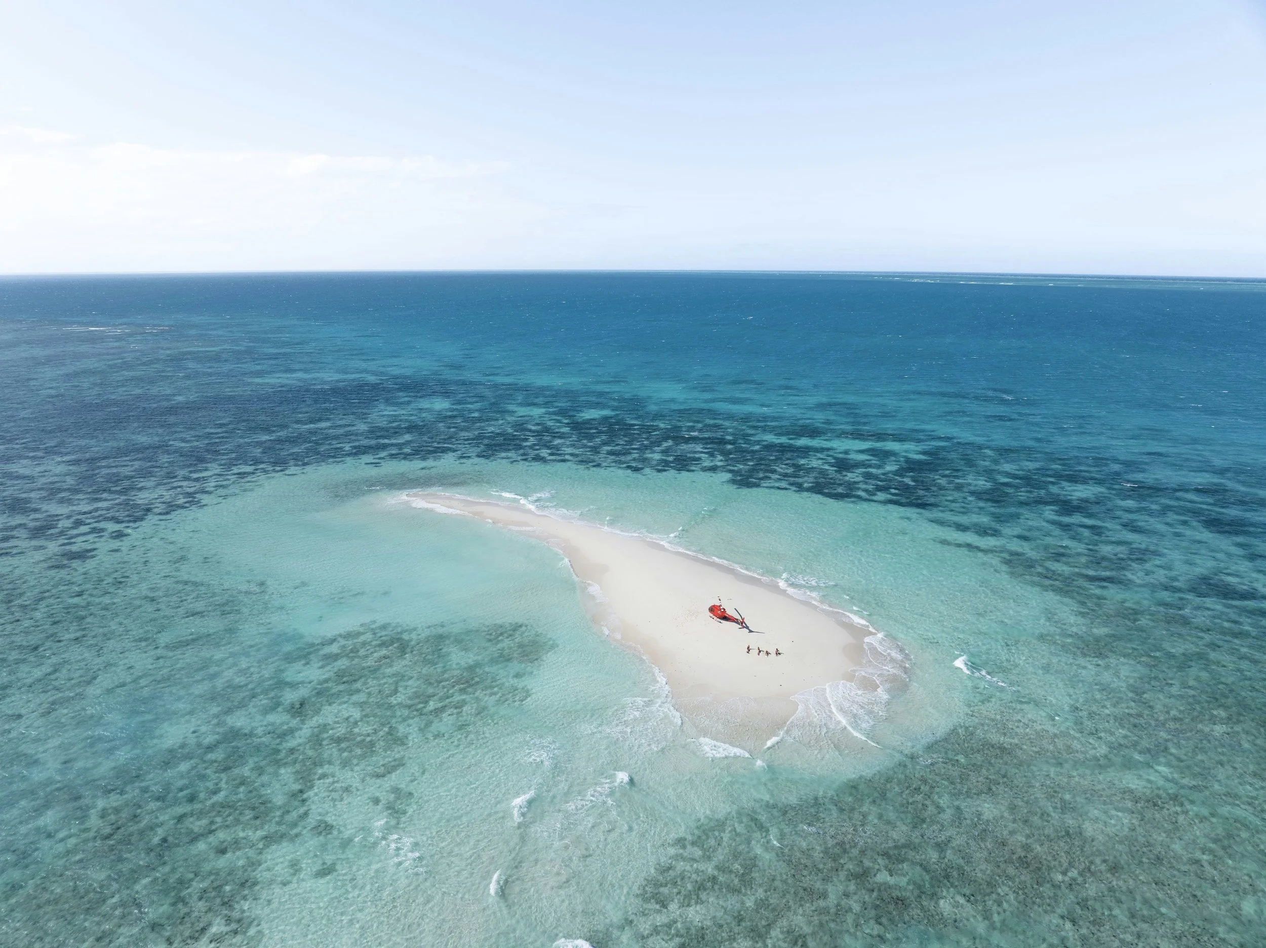 Aerial view of a small sandy island surrounded by turquoise ocean water, with a group of people on the beach and some boats nearby.