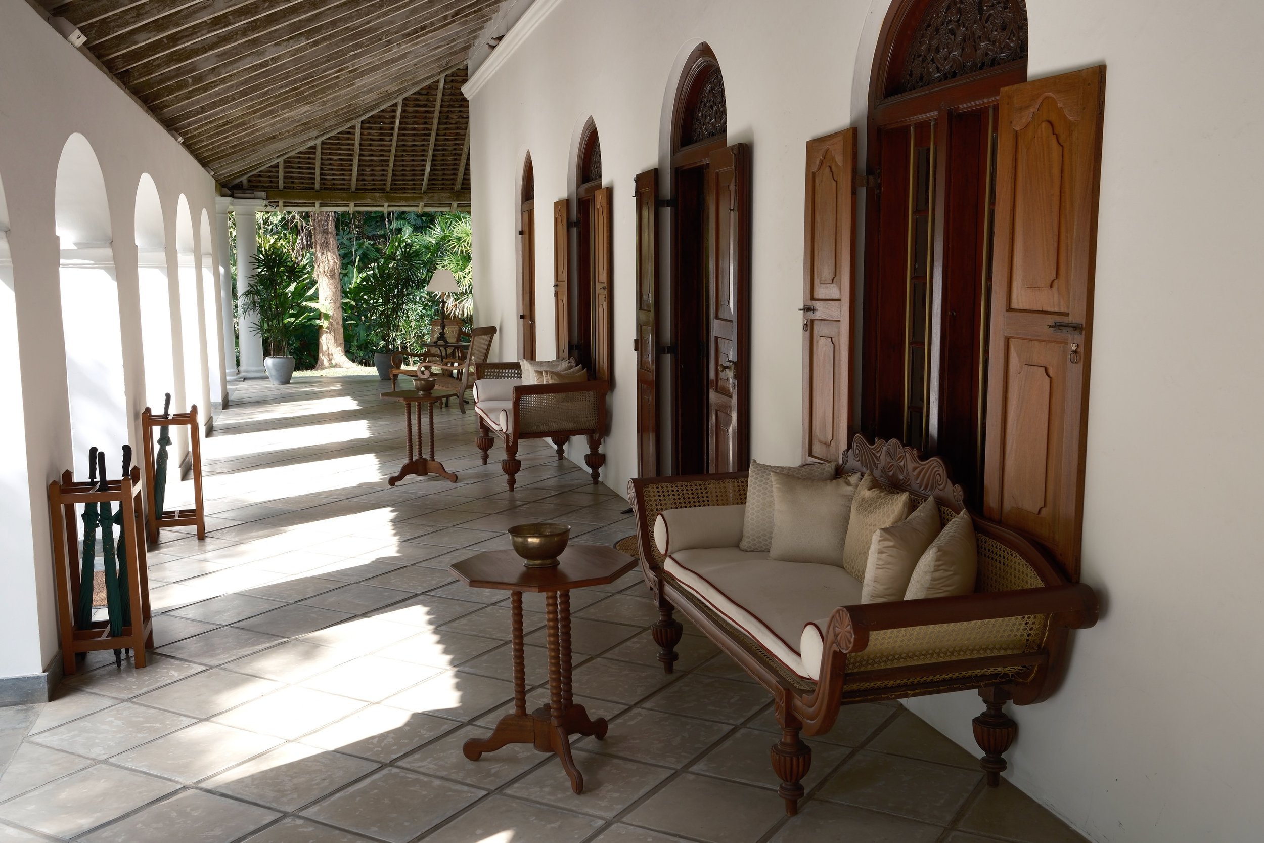 A covered porch or veranda with white walls, wooden arch windows, and wooden furniture including chairs and a sofa, with plants and trees in the background.