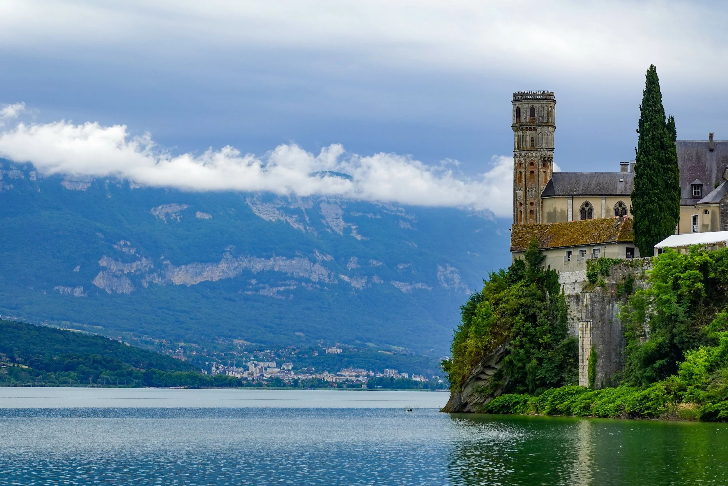 A lakeside view featuring an ancient stone tower and a church on a steep hill, with lush green vegetation, surrounded by mountains partially covered by clouds, and a calm lake in the foreground.