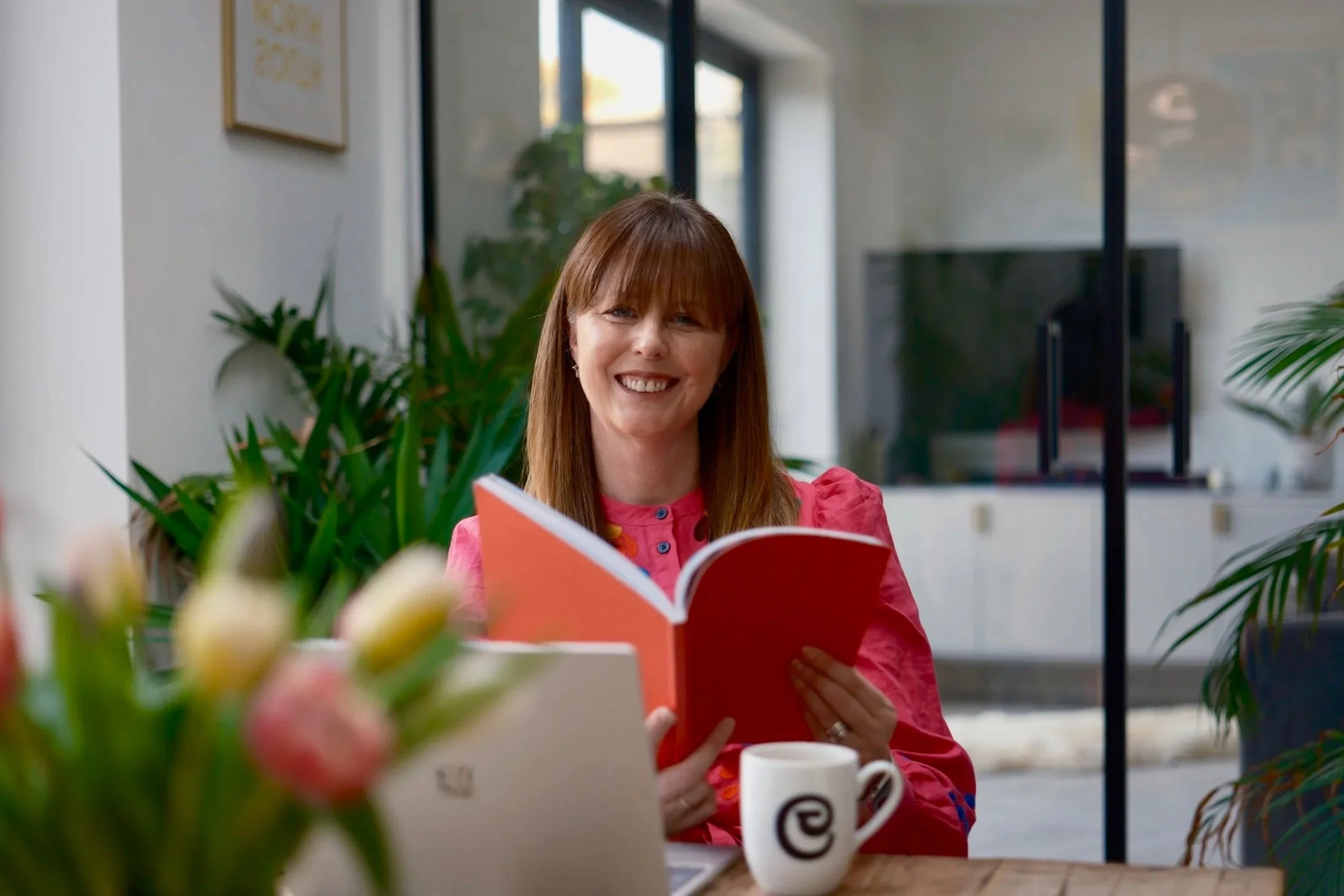 Donna Richmond Travel Councillor  in a pink blouse reading a red book at a table with coffee and plants