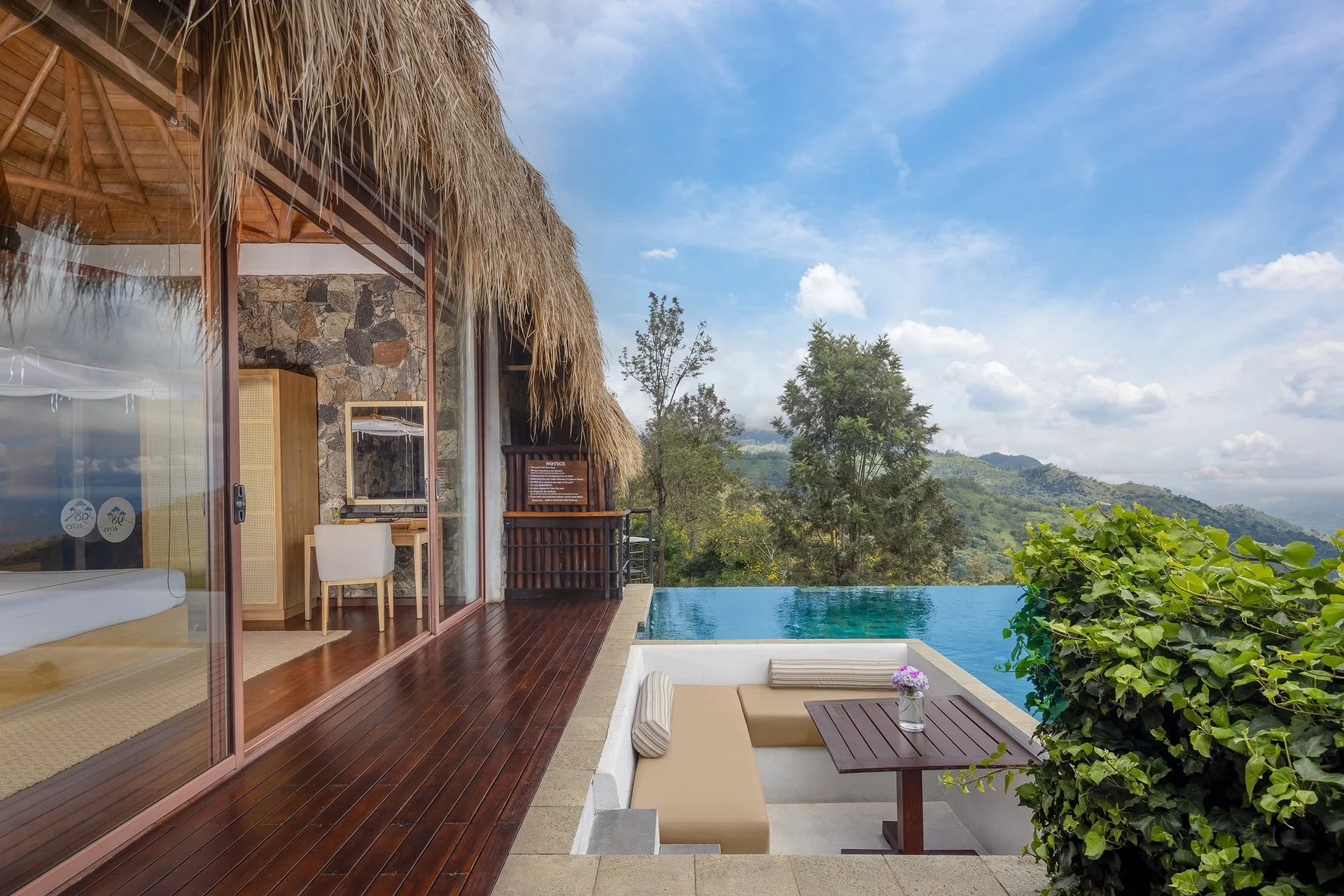 View from a balcony of a modern house with a thatched roof: glass sliding doors leading to an interior room, outdoor seating area with beige cushions and small table with flowers, a swimming pool extending to the lush green hills, and a bright blue sky with scattered clouds.