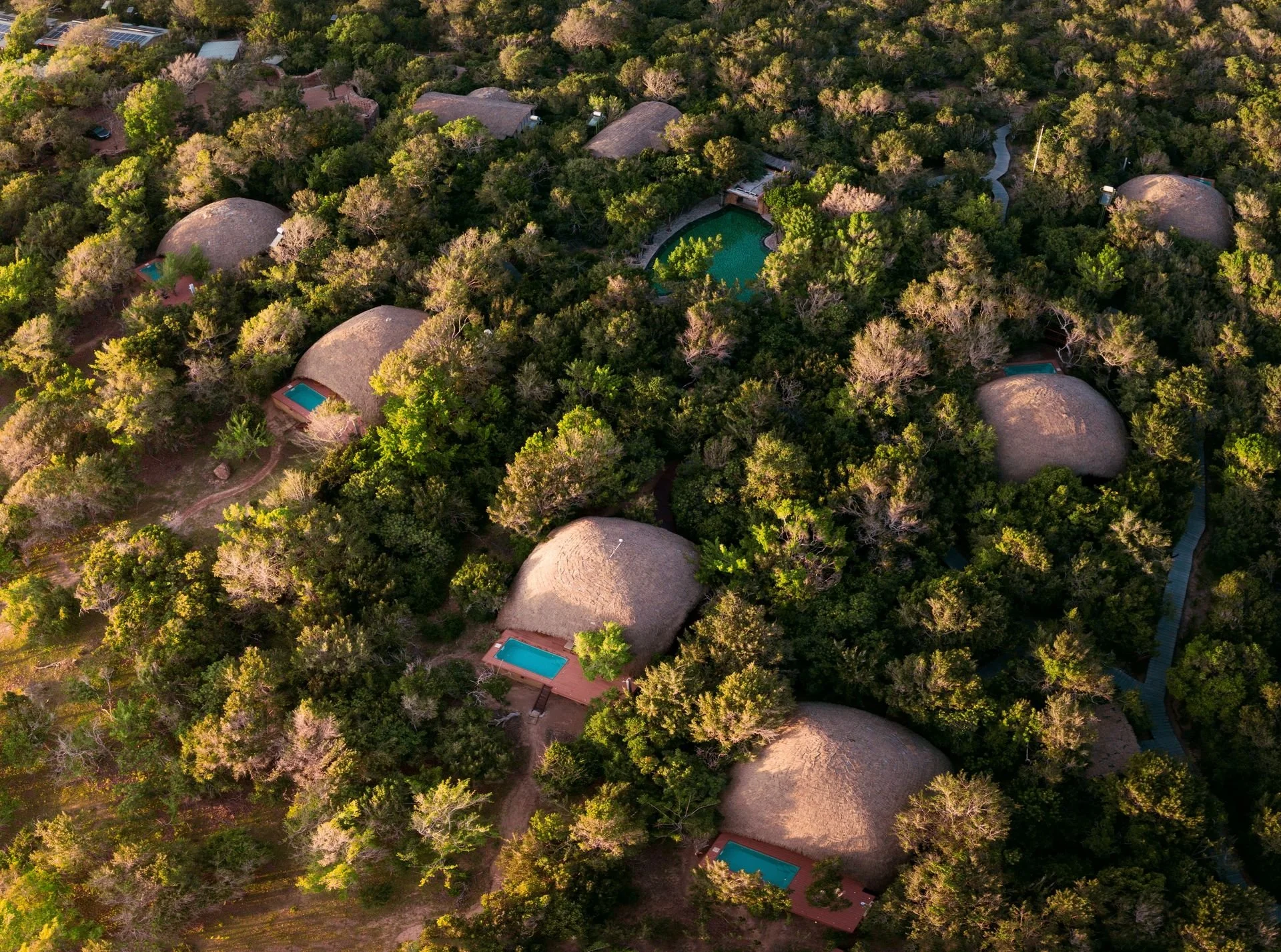 Aerial view of a resort with multiple thatched roof bungalows surrounded by dense green forest, with several swimming pools visible.