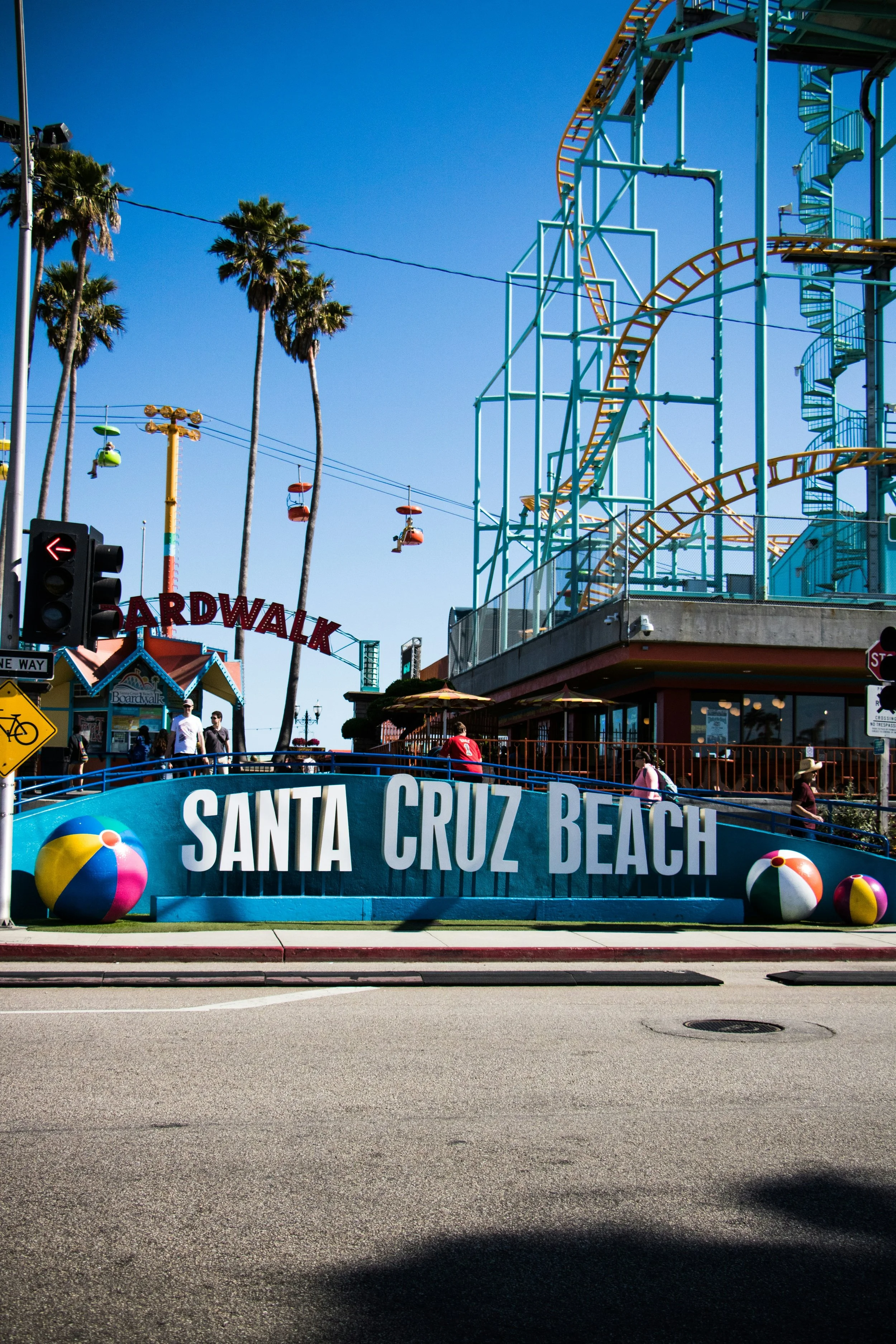 People walking along Santa Cruz Beach Boardwalk, which features a large blue sign with white text and beach balls, along with a roller coaster and palm trees in the background under a bright blue sky.