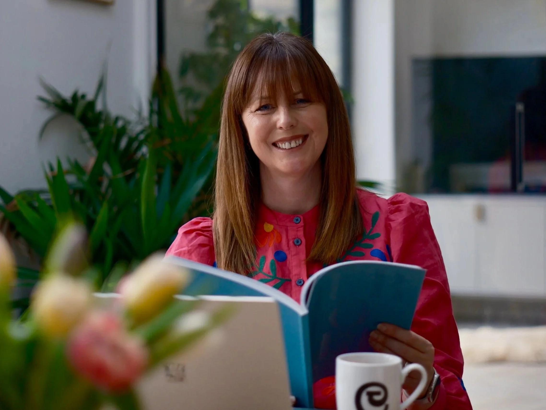 Donna Richmond in a pink embroidered blouse reading a book while holding a coffee mug at home